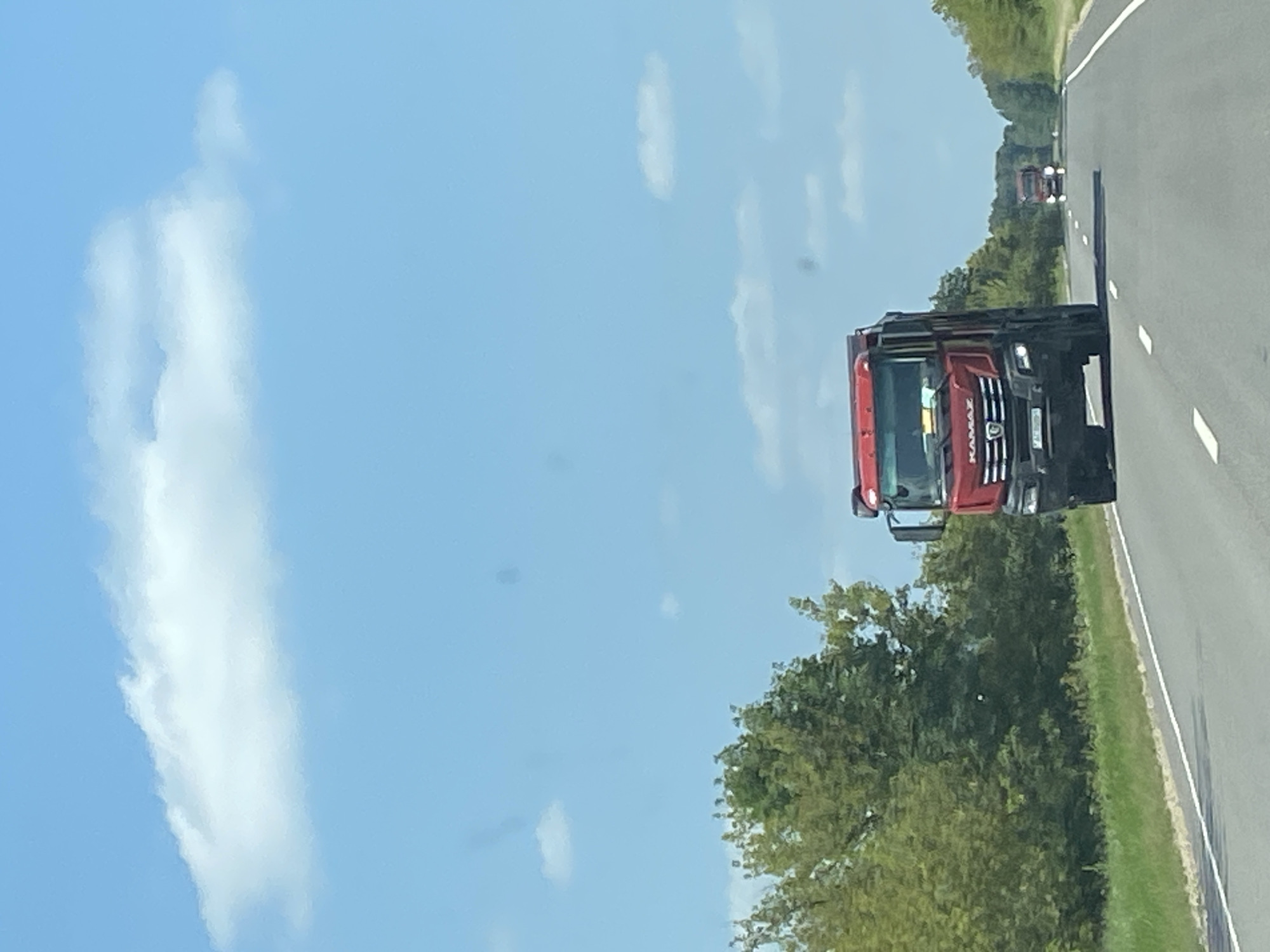 The image depicts a bright, sunny day with a clear blue sky and scattered white clouds. The foreground shows a highway with a white lane marker. A large red Kamaz truck is in the right lane, moving away from the viewer. The truck has a black bumper and grille, with a yellow sign visible through the windshield. Trees and greenery line the left side of the highway. In the distance, another red truck can be seen on the same highway. The scene appears to be in a rural or semi-rural area, consistent with the location of Матыкальскі сельскі Савет, Belarus.
