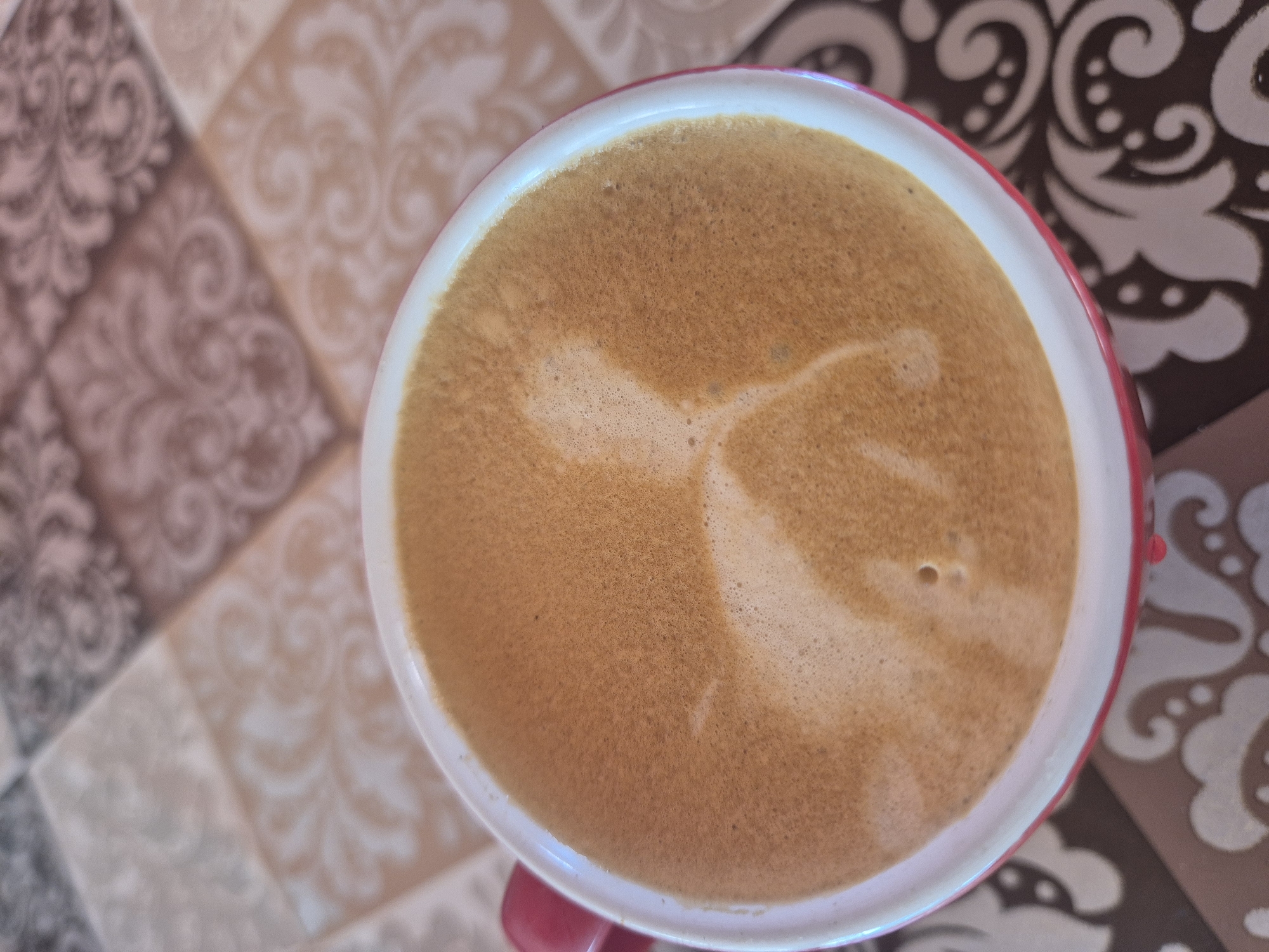 A close-up, overhead shot shows a white mug with a red rim filled with coffee, sitting on a patterned surface. The coffee has a foamy top with a light-colored design resembling a dog. The surface beneath the mug is tiled with a repeating pattern in shades of brown, cream, and grey.