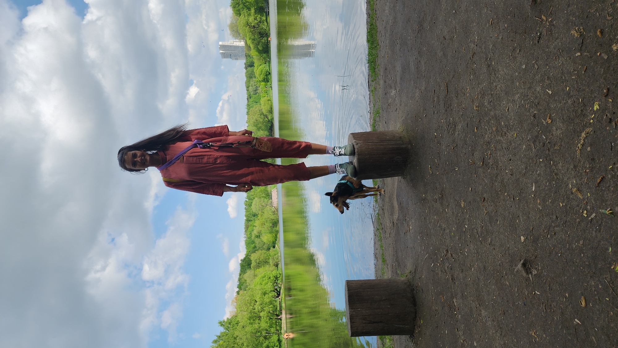 A woman stands on a tree stump beside a lake, accompanied by a small dog. The woman wears a reddish-brown jumpsuit and sneakers. A building is visible across the lake. The photograph was taken in the city of Khimki, Russia.