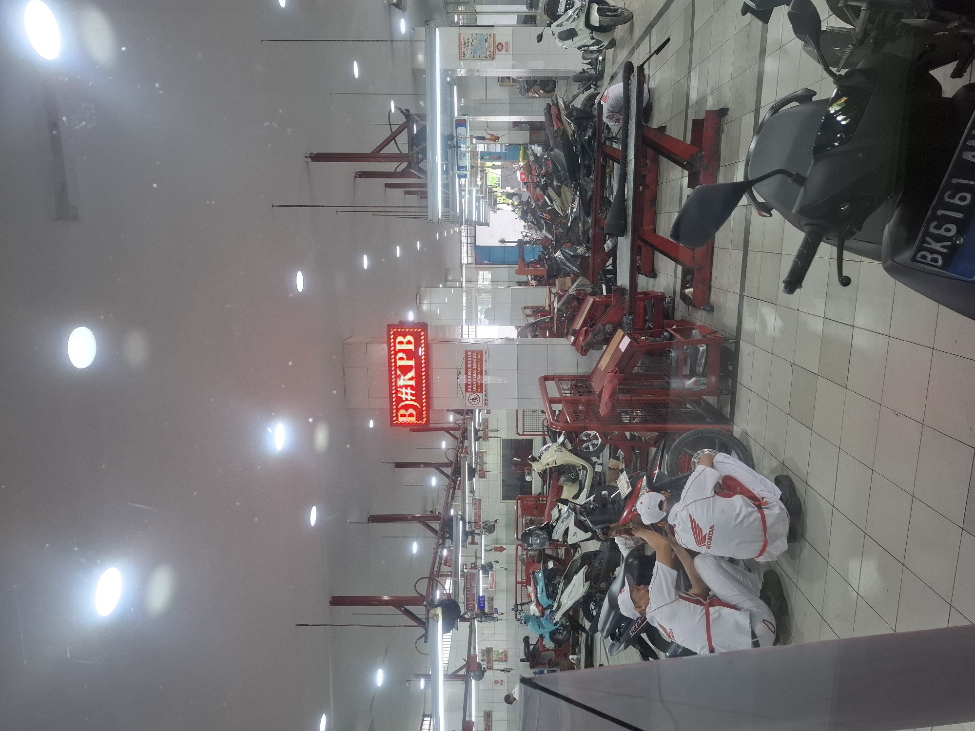 The image depicts the interior of a Honda motorcycle service center in the city of Medan, Indonesia. The environment is brightly lit by fluorescent lights on the ceiling. Several motorcycles are present, some on repair lifts. Two men in white Honda uniforms are working on a motorcycle in the foreground, one squatting and the other kneeling. In the background, more motorcycles are visible, along with tools and equipment typical of a service center. A red digital sign above some of the stalls reads "B)#KPB". A white sign with red text indicating "DILARANG MASUK AREA KERJA BENGKEL DENGAN ALAS KAKI KOTOR" is also visible. The floor is tiled with a light-colored square pattern. A black motorcycle is prominently displayed in the bottom right corner of the frame.