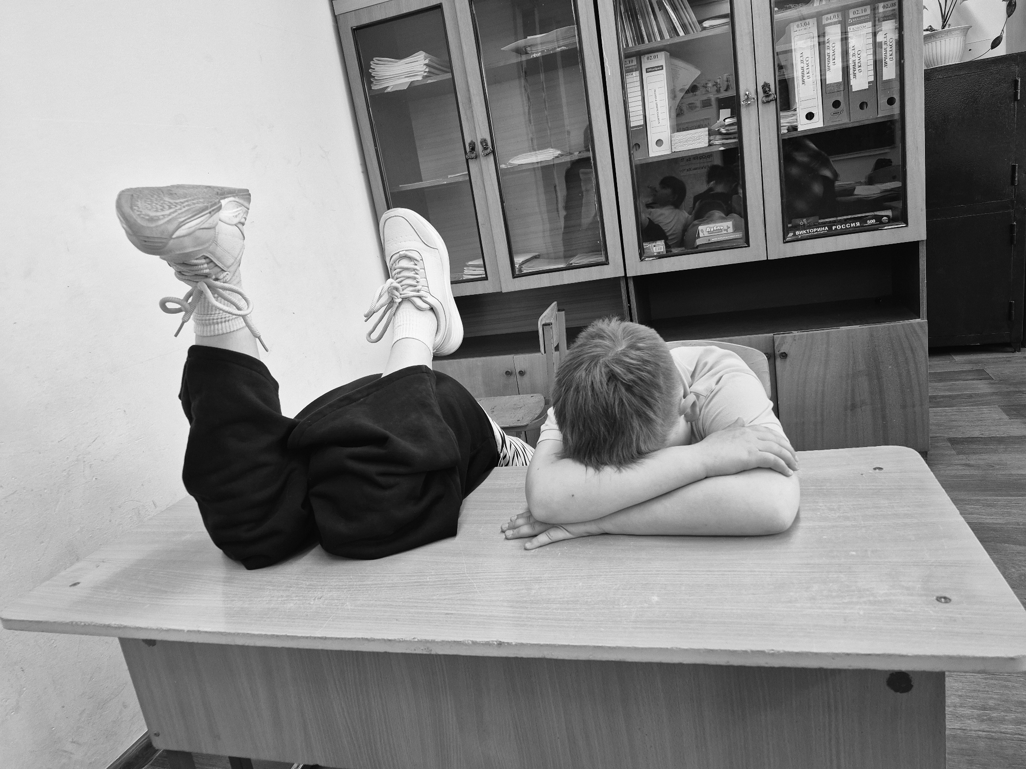 A black and white photograph depicts a boy lying face down on a wooden desk with his legs raised in the air. The boy is wearing a light-colored shirt, and his arms are crossed on the desk. He has short hair. The boy's lower half is wearing black shorts and white socks and sneakers. The shoes are facing upward, with their soles partially visible. The desk is plain and light-colored, showing some wear.
Behind the desk, there is a set of cabinets with glass doors. The cabinets contain various books and papers. There are also several binders with labels in Russian. The labels identify them as “Personal Files”. The writing on the binders indicates they are associated with different grade levels (2nd, 3rd, and 4th grade). The environment suggests a classroom or office setting in a school. The cabinet also contains a book that says “VIKTORINA ROSSIIA 500,” which translates to “Quiz Russia 500.”
The location is the city of Chaikovsky, Russia, indicated by the Cyrillic text on the binders. The rest of the room is plain, with a white wall visible in the background. Part of a dark cabinet or storage unit is visible at the right of the image.