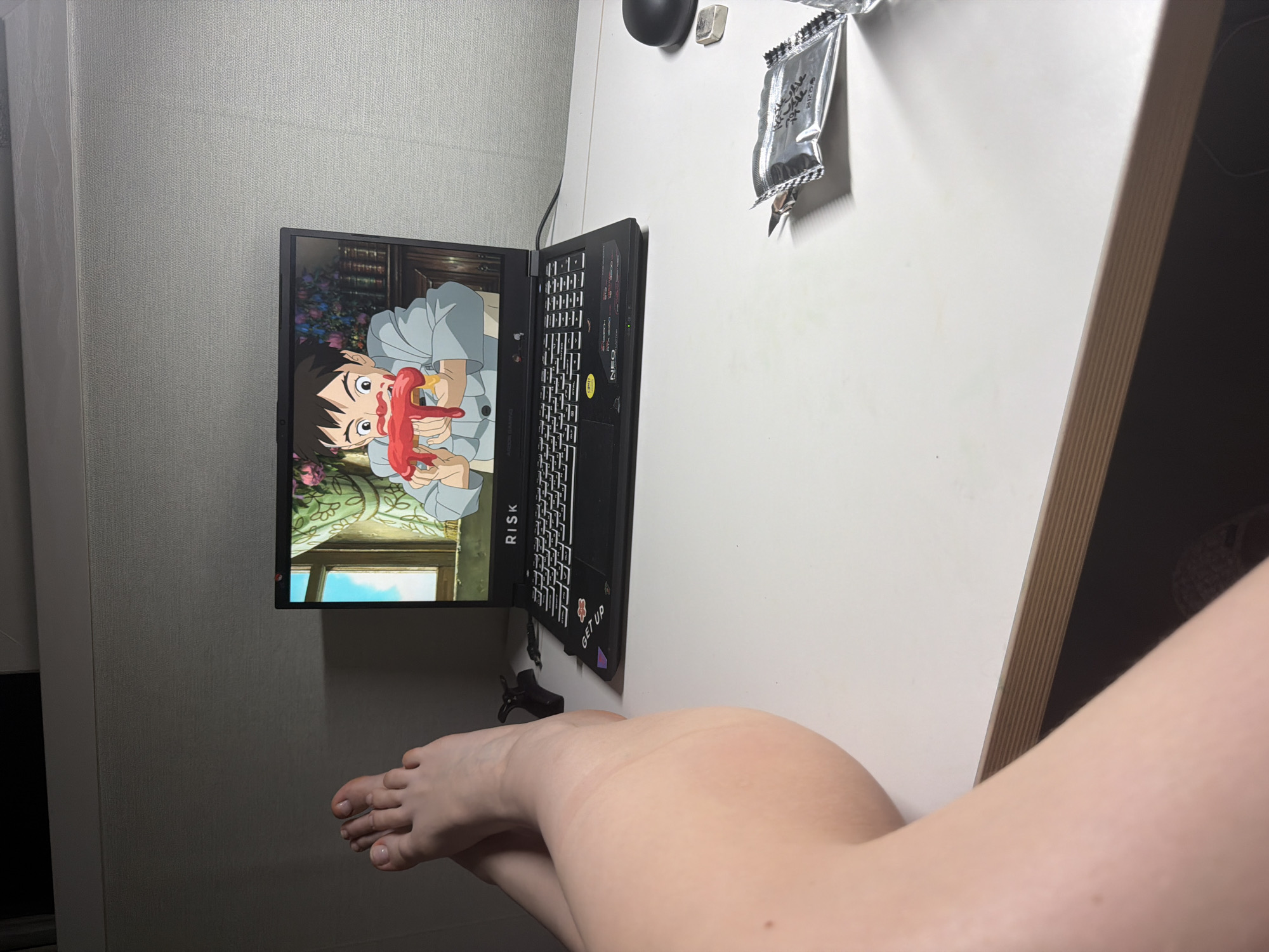 A first-person perspective shows a person's bare legs and feet crossed in the foreground, positioned on a white desk. To the right of the legs, a black laptop is open, displaying an anime scene of a young boy eating a hot dog. The laptop's screen shows the character from the anime "Castle in the Sky." The desk also has a black computer mouse to the right of the laptop, and a small silver object. In the middle right of the desk, a silver packet with Korean text is partially open. The background consists of a textured gray wall. The overall setting appears to be a room in Dongducheon-si, South Korea.