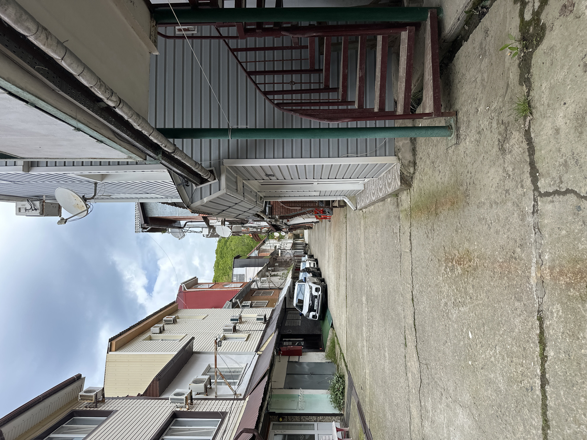 The image depicts a narrow street in Novomikhaylovsky, Russia, lined with multi-story residential buildings of varying designs and colors. Several air conditioning units are mounted on the building facades. A row of parked cars occupies the center of the street. Concrete sidewalks flank the buildings. Overhead, a network of pipes and wires runs along the buildings, with satellite dishes also visible. A metal staircase is visible attached to one of the buildings. The sky is overcast.
