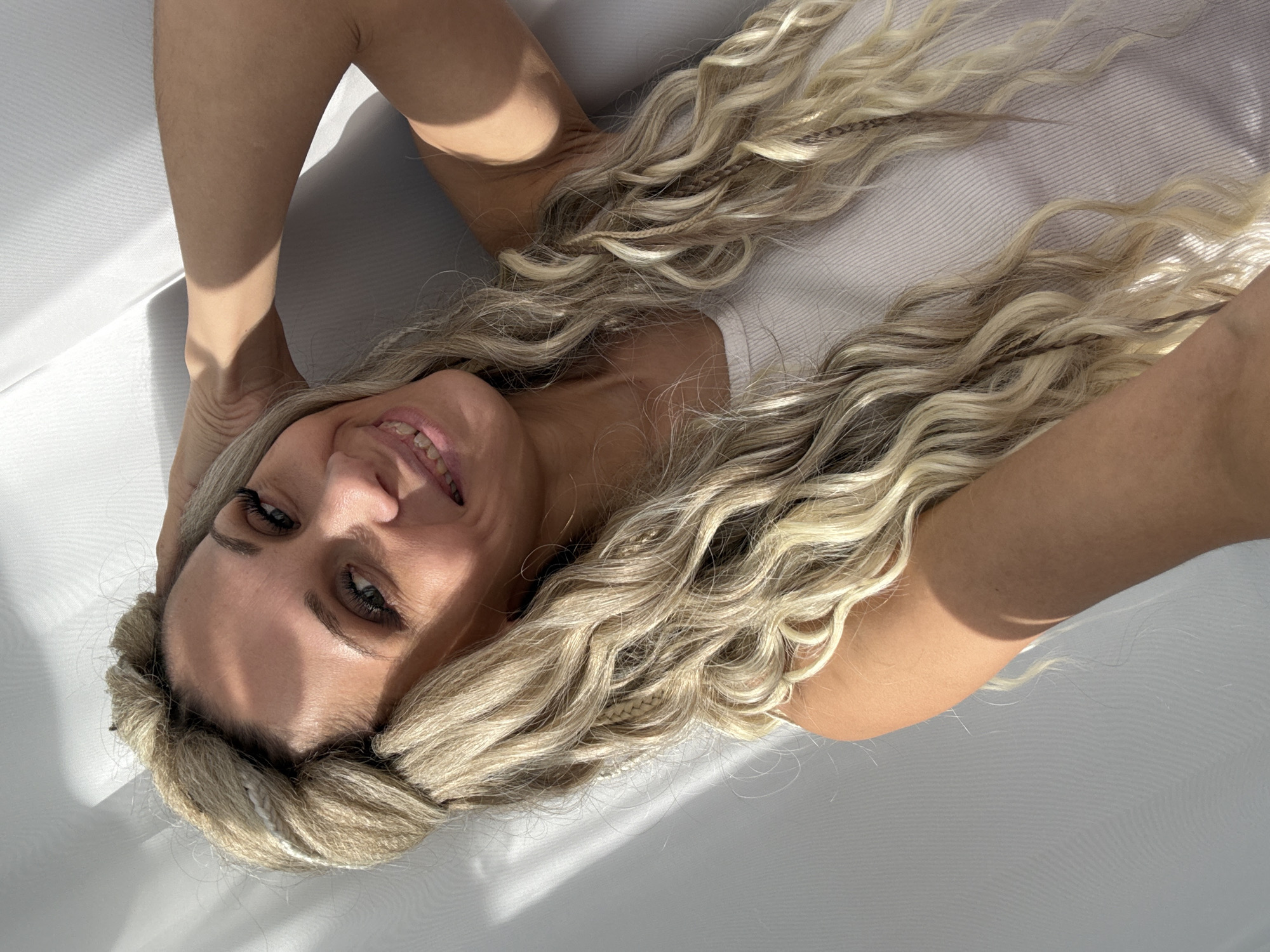 A woman with long, flowing blonde hair is depicted lying down, looking up at the camera with a cheerful, relaxed smile. Her head is towards the bottom left of the frame, and her arms are slightly raised, framing her face. Her hair, which is a mix of wavy textures and thin braids, spreads across a plain, light-colored surface, possibly a bed or couch, filling much of the background.
The woman has fair skin, dark eyebrows, and dark, defined eyelashes. Her lips are parted in a wide smile, revealing her teeth. She appears to be wearing a light-colored, possibly white or off-white, top. The scene is illuminated by bright, directional light, likely sunlight, which casts distinct, geometric shadows across the white background and parts of her skin, suggesting an indoor setting with light coming through a window or opening. The shadows create clear lines and patches of darker tones against the brighter areas.
The image is taken from a top-down perspective, with the woman positioned on her back. The overall mood is positive and serene, emphasized by her pleasant expression and the soft, natural lighting. There is no visible text in the image. The location is an unknown indoor setting, and the time of day appears to be sunny daylight.