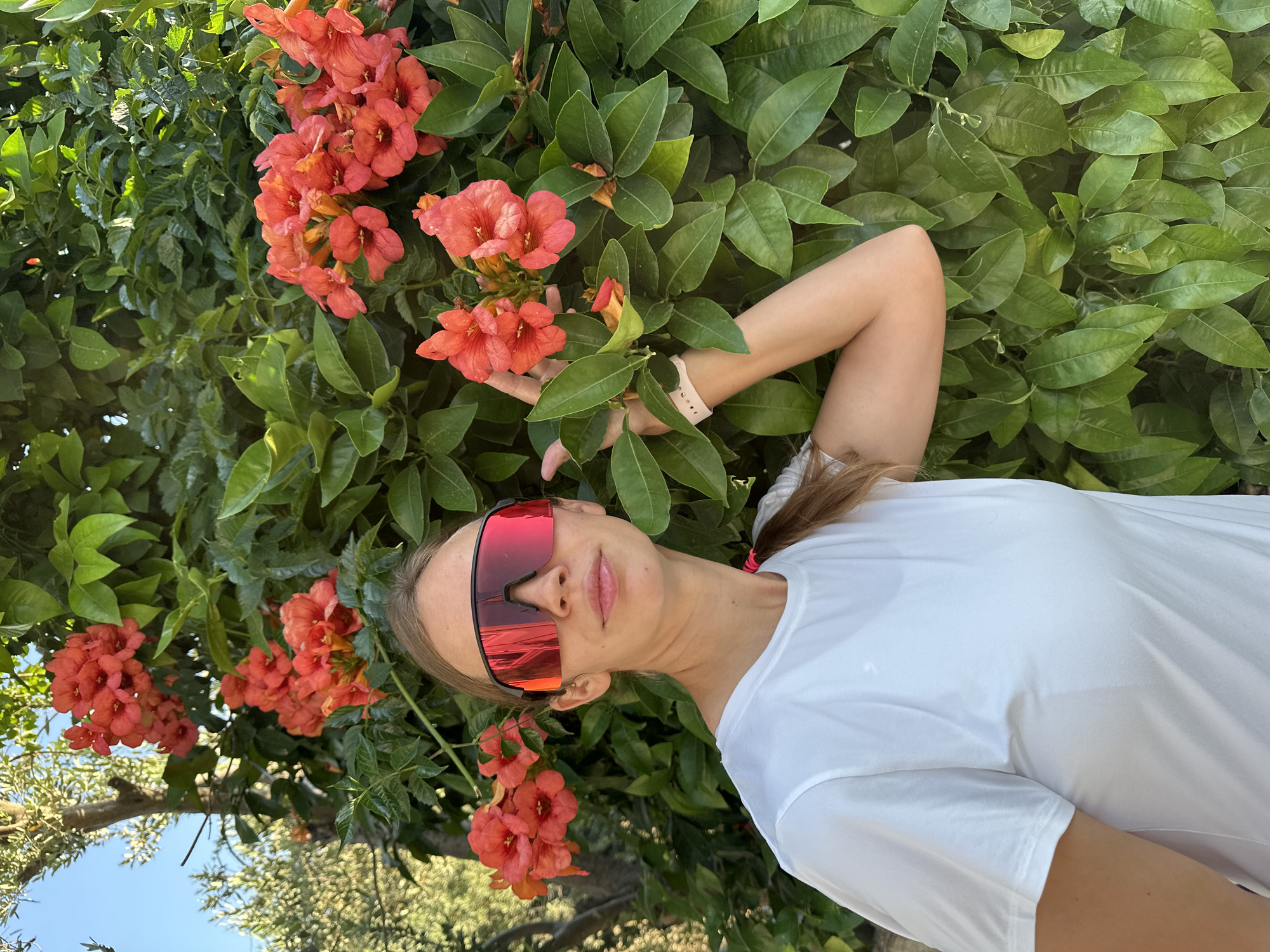 A female individual stands outdoors in a bright daylight scene within the city of Fethiye, Turkey. She has light brown hair, partially visible in a ponytail secured with a pink hair tie, and wears a plain white short-sleeved t-shirt. Her eyes are obscured by large, wrap-around, red mirrored sunglasses with black frames. Her lips are slightly parted in a subtle smile. Her left hand is raised, gently touching a cluster of vibrant orange-red, trumpet-shaped flowers. A light-colored, possibly white or light pink, smartwatch or fitness tracker is visible on her left wrist.
The flowers are part of a dense, leafy vine or shrub with numerous dark green, oval-shaped leaves that forms the immediate background. Beyond this plant, additional green foliage is visible, including the trunk and leaves of what appears to be an olive tree in the upper left portion of the frame. A clear blue sky is visible above the trees and foliage in the upper left. The sunlight appears strong, illuminating the scene and creating highlights on the leaves and the woman's clothing.