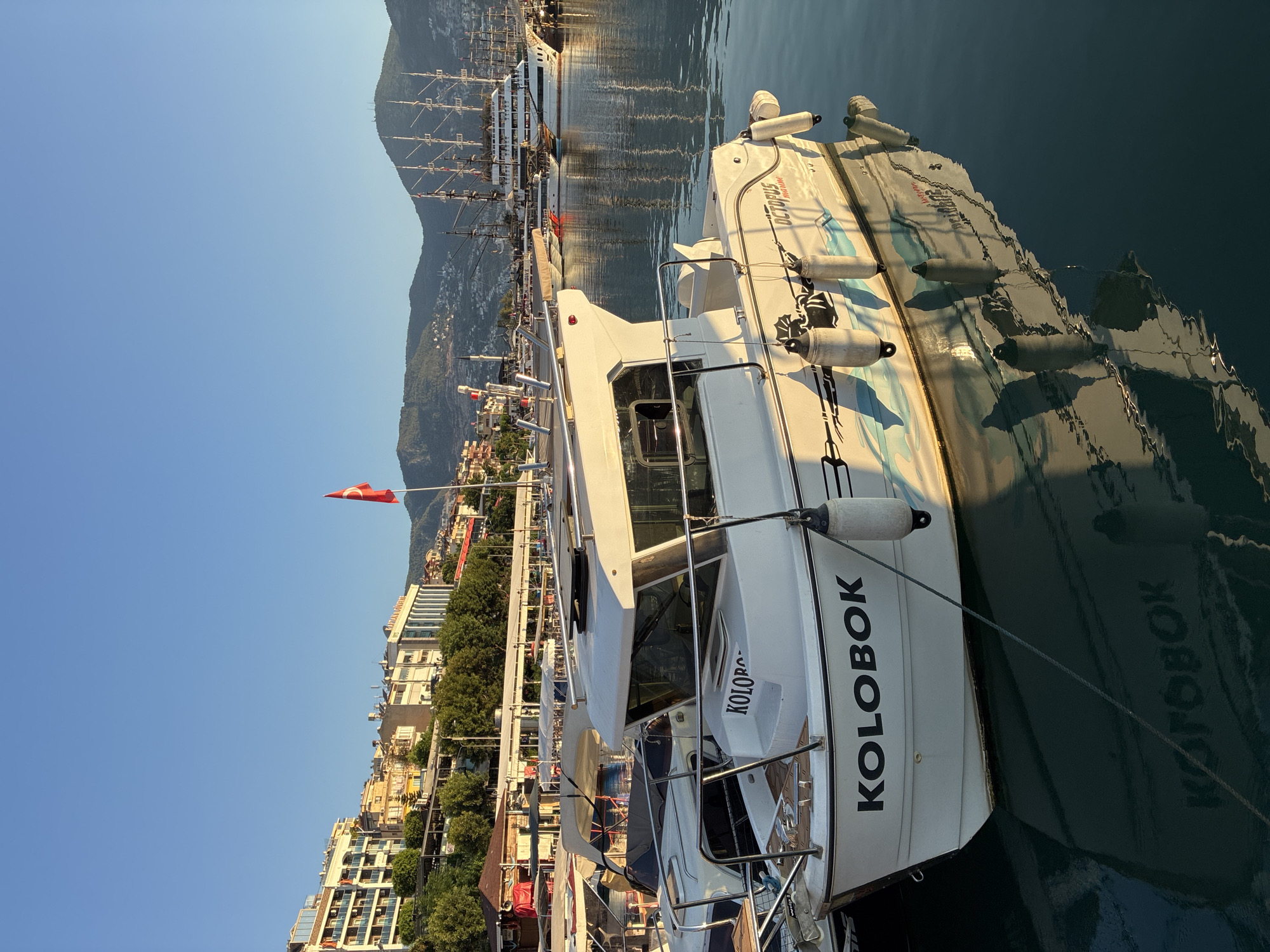 The image depicts a harbor in Alanya, Turkey, bathed in the warm light of a setting or rising sun. In the foreground, a white motorboat named "KOLOBOK" is prominently displayed, tethered to a dock. Its reflection is clearly visible in the calm, dark blue water. To the left of the "KOLOBOK," other boats are partially visible, also docked.
In the mid-ground, the Alanya Marina is seen with numerous other vessels and a promenade lined with restaurants and shops. A large, multi-masted sailing ship, possibly a replica, is anchored further out in the harbor. The backdrop consists of the cityscape of Alanya, characterized by numerous buildings cascading down a hillside, leading up to lush green mountains. A Turkish flag flies from a flagpole on the hillside. The sky is a clear, vibrant blue, indicating fair weather. No people are visible in the image.
