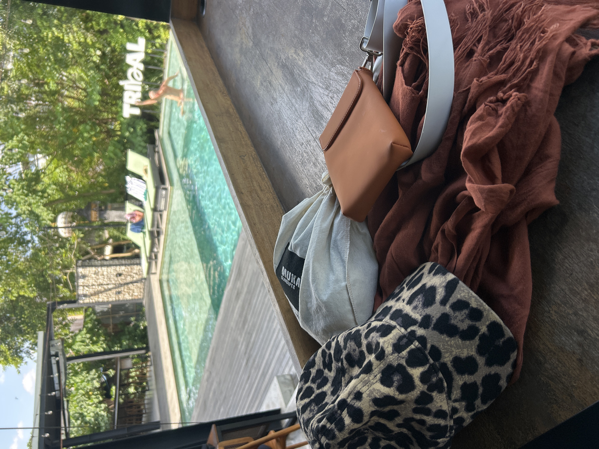 The image presents a vibrant, tilted view overlooking a swimming pool area, with personal items occupying the immediate foreground.
On a dark, textured wooden surface, likely a railing or table, several objects are neatly arranged. These include a small, light brown (tan/camel) faux leather pouch with a flap closure, suspended by a white strap. Adjacent to it is a crumpled, rusty-red or burnt orange fringed scarf or textile. Also present are a light grey drawstring bag featuring the black text "MUKA CONCEPTS," and a striking leopard print bucket hat in black and beige/tan.
Beyond the foreground items, a clear turquoise swimming pool stretches out. A person is visible in the water, seemingly standing or moving near the pool's edge. Further back, another individual is seen relaxing on a green lounge chair by the poolside. A large white sign with the bold, capitalized word "TRIBAL" is prominently displayed, partially submerged in or situated at the edge of the pool. The entire background is rich with lush green trees and tropical foliage, indicating a natural and serene outdoor setting. Some architectural elements with stone-like textures are subtly visible through the greenery.
The scene captures a sunny daytime, suggesting leisure and relaxation in a tropical environment. The visible text includes "TRIBAL" on the pool sign and "MUKA CONCEPTS" on the drawstring bag. The location, as provided, is Mengwi, Indonesia.