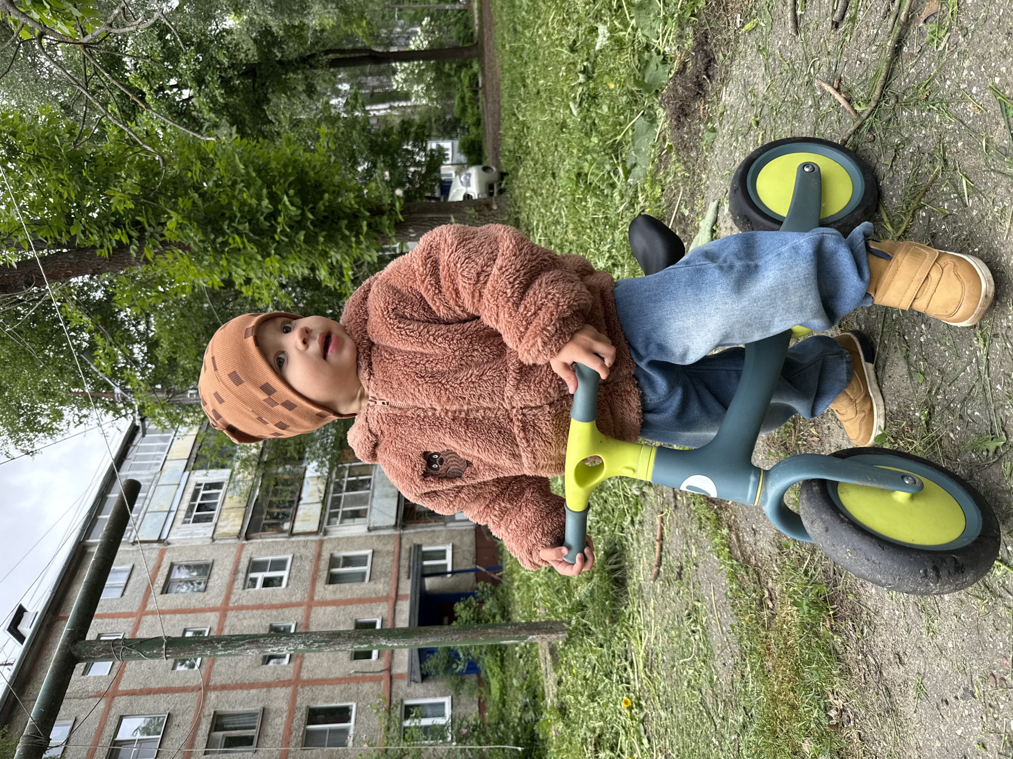 A young child wearing a brown hat, brown fluffy jacket with an animal emblem, blue jeans, and tan shoes sits on a blue and lime green tricycle on a patch of grass and dirt. The backdrop includes a large apartment building with many windows, trees, and other vegetation. The location is suggested to be Rusanovo, Russia.
