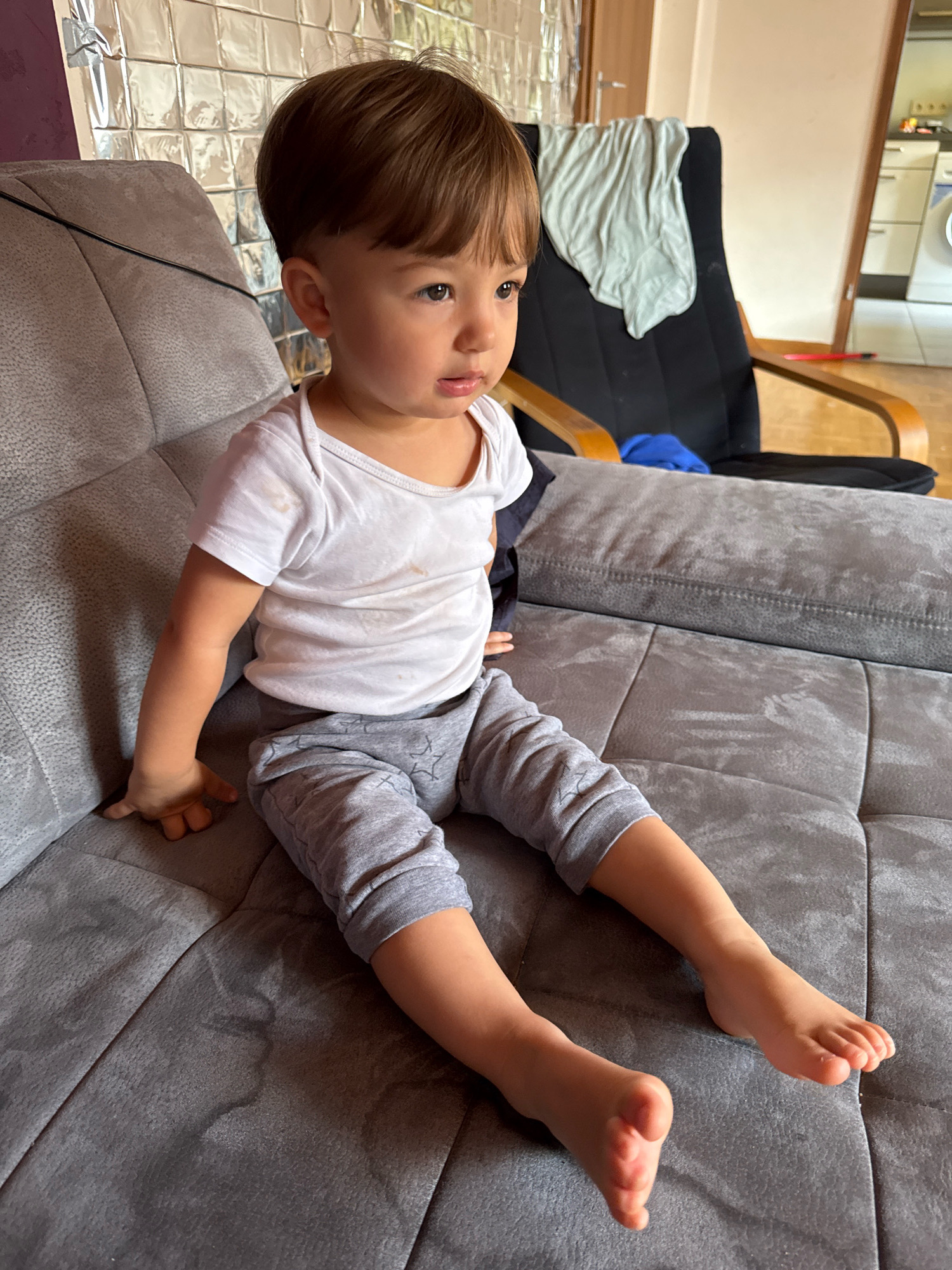 A young child, appearing to be two or three years old, sits on a gray sofa in what appears to be a residential setting. The child has brown hair and is wearing a white t-shirt and gray sweatpants with a star pattern. The child's left arm is bent, with their hand resting on the sofa, and their legs are extended forward, with their bare feet visible. The sofa has a textured, tufted surface. In the background, a black chair with wooden armrests is visible, draped with a light-colored cloth. Further in the background, a doorway leads to what seems to be a kitchen area with white cabinets and a countertop. The overall scene suggests a casual indoor environment. The location is identified as Überherrn, Germany.