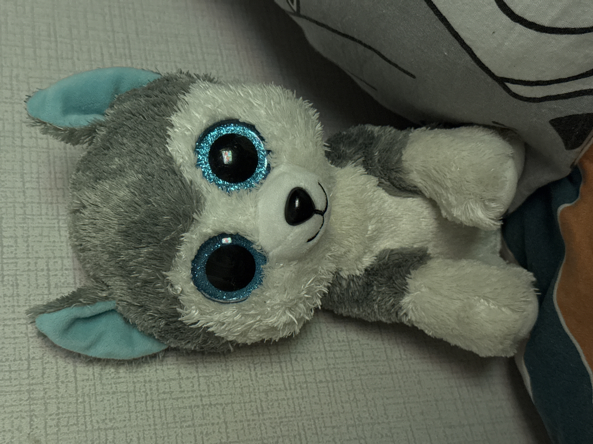 A stuffed husky dog toy with large blue glittery eyes sits in front of a textured gray wall. The toy has gray and white fur and two light blue ears. A pillow with a black outline drawing of a face is to the right of the toy. The toy is positioned on a surface with a striped pattern of dark blue, orange, and white. The scene is indoors.