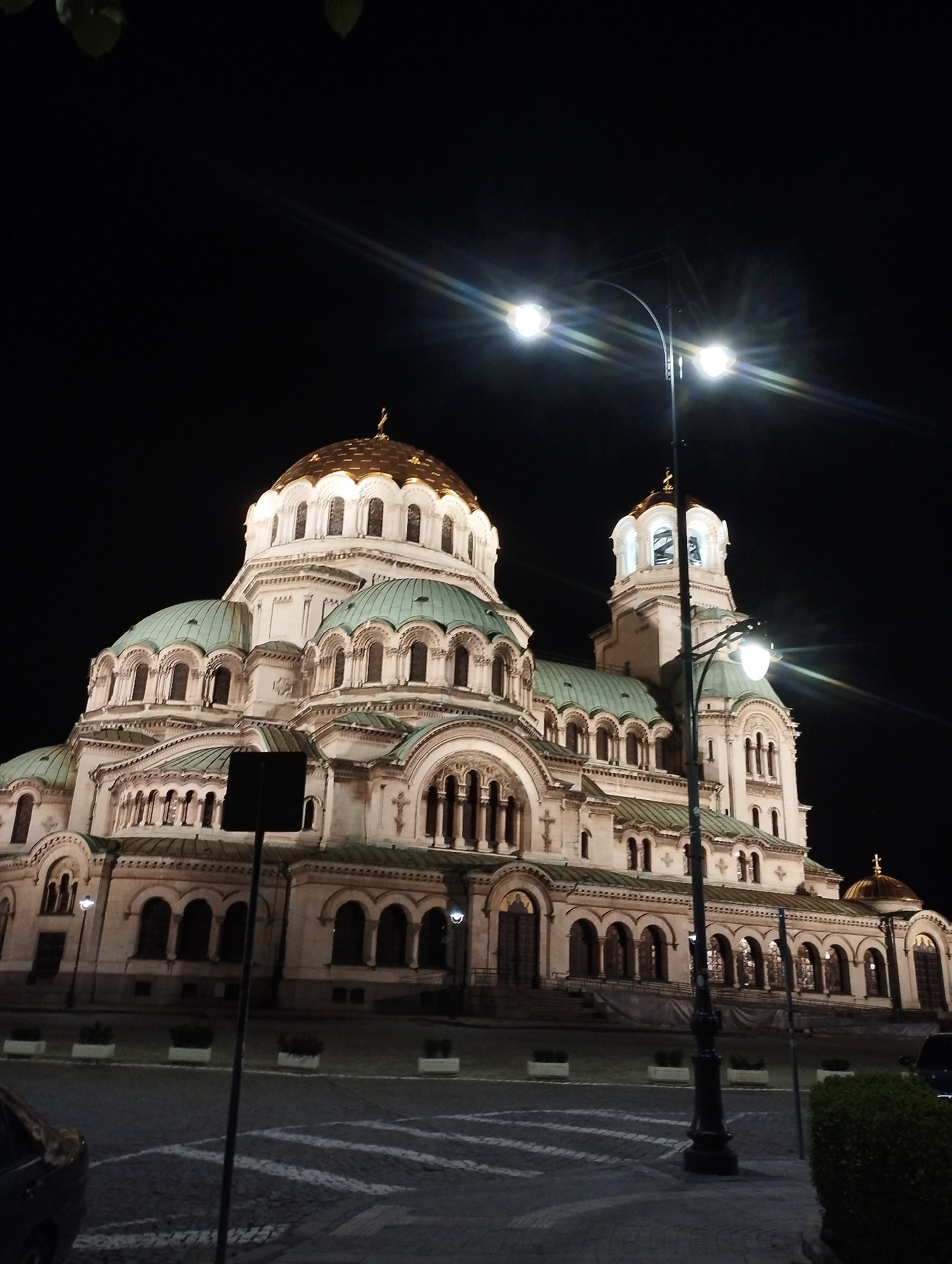 The image displays a grand, ornate cathedral-like building at night, illuminated against a deep black sky. The structure is characterized by multiple domes, including a large central golden dome and several smaller, teal-green domes. Its facade is richly detailed with numerous arched windows, decorative columns, and intricate stone or stucco work, all brightly lit by external lights.
A prominent dark streetlight with two glowing lamps stands in the foreground, slightly to the right of the center, casting strong light and noticeable lens flares across the dark sky. Below the building, patterned pavement, possibly cobblestone, is visible, along with parts of a dark car on the far left and a dark bush in the bottom right corner. Another dark pole or signpost is present in the mid-ground.
The time of day is clearly nighttime, evidenced by the black sky and the reliance on artificial lighting to highlight the building's architectural features. There are no people visible, and no specific activity or event is taking place, presenting a static, majestic view of the illuminated edifice. No visible text can be discerned in the image.