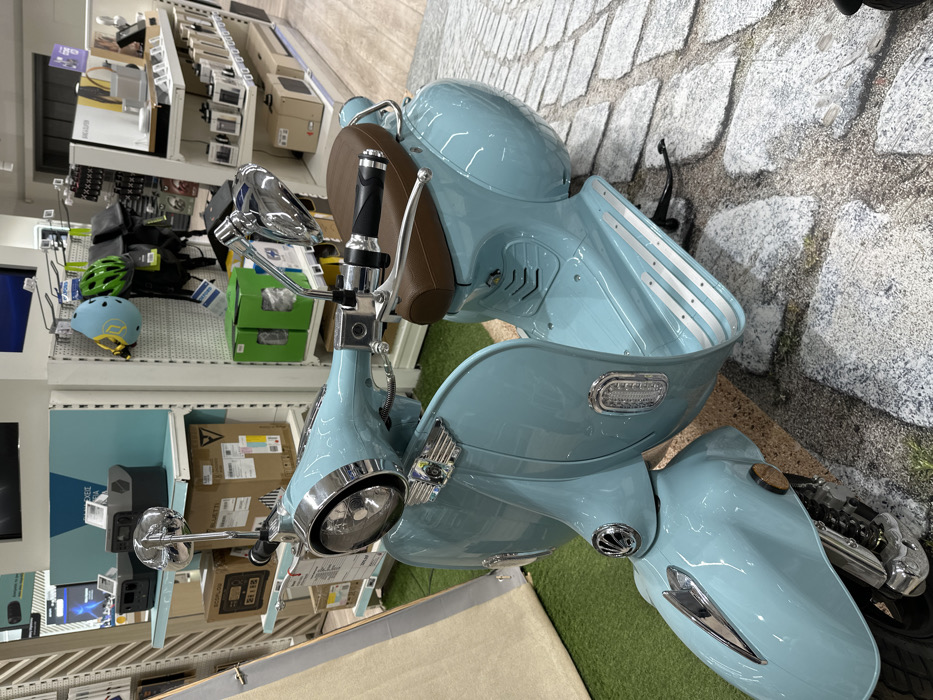 This media file captures a light blue, vintage-style scooter displayed in what appears to be a retail store or showroom in Metamorfosi, Greece. The scooter is the primary subject, angled diagonally and dominating the foreground and midground of the image. It features a shiny, light blue body, a brown leather-textured seat, chrome handlebars with black grips, chrome mirrors, a prominent chrome-bezeled headlight, and turn signals. The footrest area includes silver, ribbed anti-slip strips. Visible elements of the engine and front suspension are also seen near the front wheel.
The scooter is positioned on a display surface featuring artificial green turf and a cobblestone-patterned mat, creating an outdoor ambiance indoors. In the background, white metal shelving units are packed with various products. These include outdoor/sports gear such as a bright green helmet and a light blue helmet with yellow accents (possibly a "C" or "G" logo), as well as black bags. Other shelves display electronics, including boxed items and what appears to be an ECOFLOW portable power station. The store lighting is bright and artificial, suggesting an indoor setting. No people are visible, indicating the scooter is stationary and on display. The overall scene suggests a vibrant retail environment showcasing products for sale.