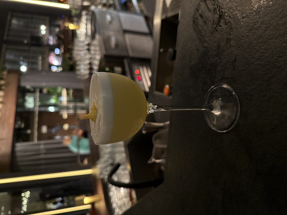 A stemmed coupe glass containing a yellow liquid topped with a white foam layer and garnished with a small, yellow, pointed element stands centered on a dark, textured bar counter. To the immediate left in the mid-ground, a black gooseneck faucet extends over a dark sink basin. To the right in the mid-ground, a row of inverted clear glasses is visible on a rack above stainless steel bar equipment featuring two red indicator lights. The background is out of focus, showing blurred architectural details, ambient lighting, and a person in a light blue shirt partially visible on the left. The scene depicts an indoor bar environment within the city of Varna, Bulgaria.
