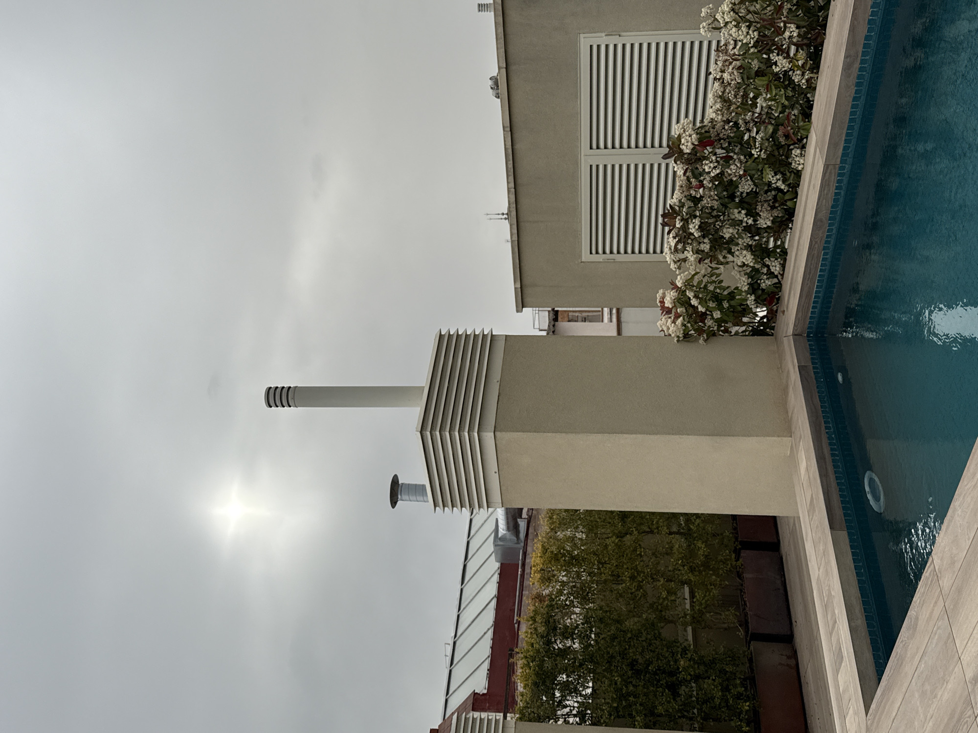 A horizontal outdoor shot features a section of a building beside a swimming pool on an overcast day, possibly in Barcelona, Spain. The building, in beige tones, includes a window with white louvers and a vertical ventilation shaft with a smaller cylindrical chimney. A bush with white flowers is planted next to the building. Part of a swimming pool fills the right side of the shot, surrounded by light-colored tiles. Below the main building, a partial view reveals another building with a glass roof and red walls, along with a dense green bush. The sky is a uniform light gray, with the sun partially visible.