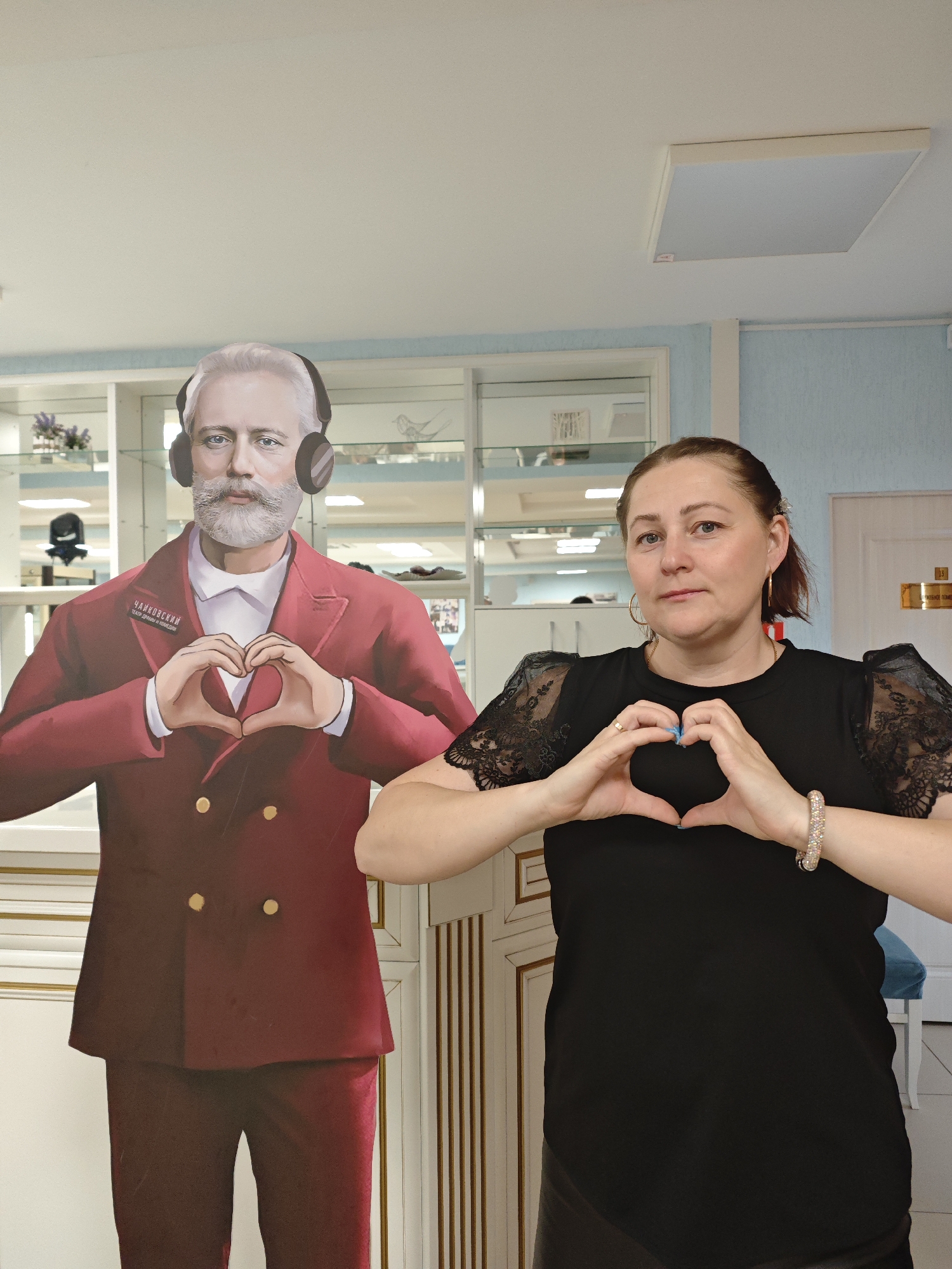A medium shot captures a woman standing next to a life-size cutout of Pyotr Ilyich Tchaikovsky in what appears to be a room with display cases. The woman and the cutout are both making heart shapes with their hands, positioned in front of their chests.
The woman, with fair skin and brown hair pulled back, wears a black top with lace sleeves, gold hoop earrings, and a sparkly bracelet on her right wrist. Her fingernails are painted a light blue color.
The Tchaikovsky cutout is wearing headphones and a red suit. A badge on his lapel reads "Чайковский," identifying him.
Behind the figures, there is a light blue wall and white cabinets or display cases containing various items. A white square light fixture is visible on the ceiling. A doorway is visible to the right of the woman. The setting suggests this photo was taken in an area related to Tchaikovsky, likely in the city of Chaikovsky, Russia, given the subject of the cutout.