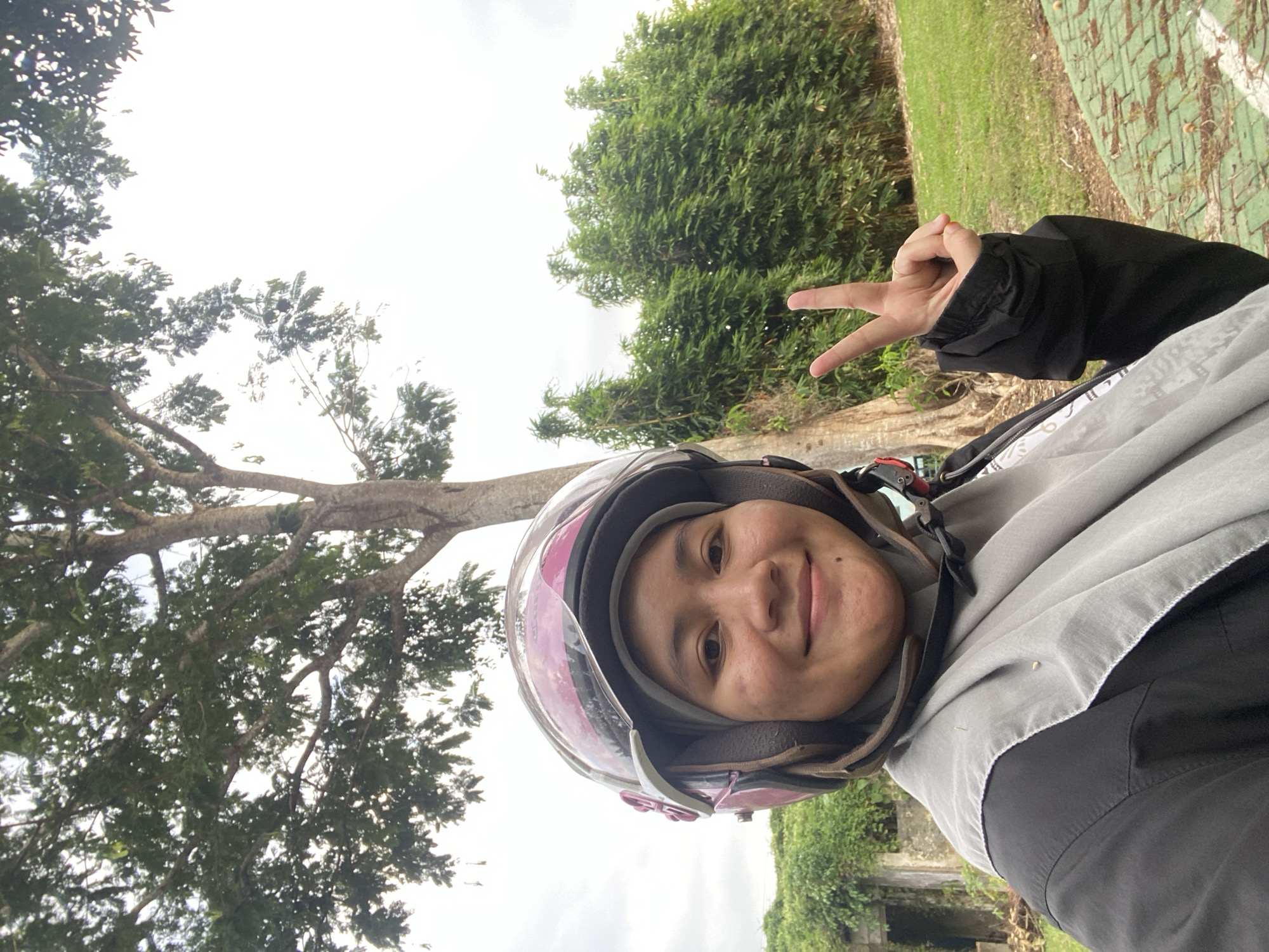 A woman wearing a hijab and a pink and brown helmet is taking a selfie outdoors. She is smiling and making a peace sign with her right hand. She is wearing a black jacket over a grey scarf. Behind her, there is a large tree, bamboo plants, and a cloudy sky. The ground is covered in grass and a green brick path. The scene appears to be in a park or outdoor recreational area. Based on the presence of these elements, the location is likely in Banjarbaru, Indonesia.