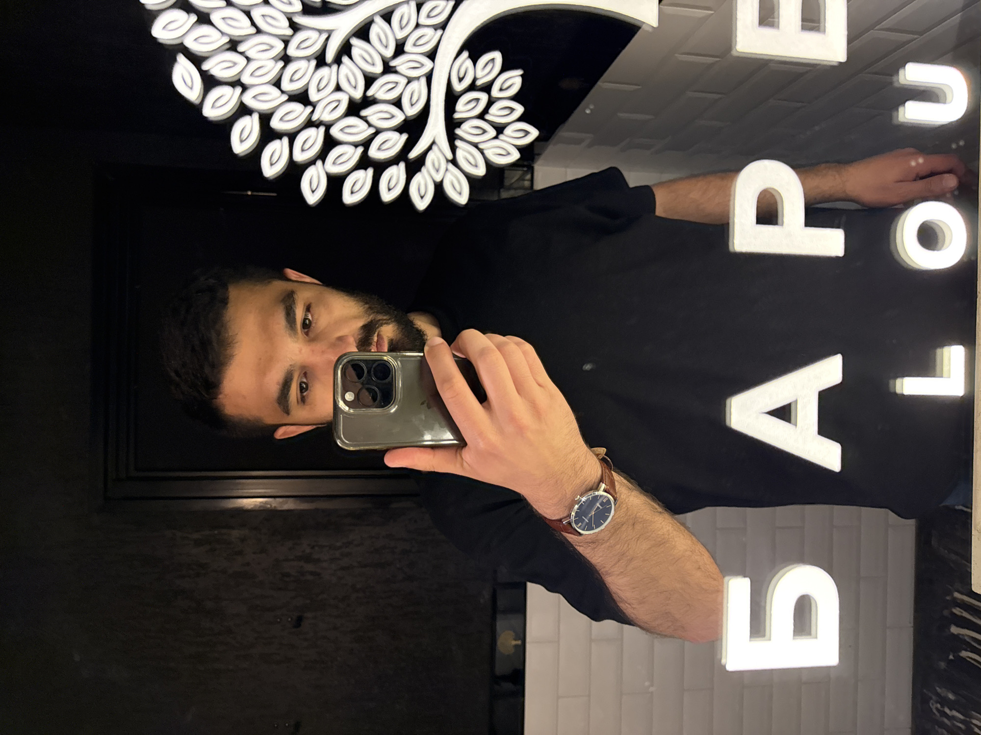 A person is taking a selfie in a mirror. The mirror's decor includes a white tree design at the top and vertically stacked letters (B, A, R, P, O, U) on the right side. The person has dark hair and a beard, and is wearing a black t-shirt and a watch with a brown band. The background behind the mirror appears to be dark. The person is using a smartphone with multiple lenses.