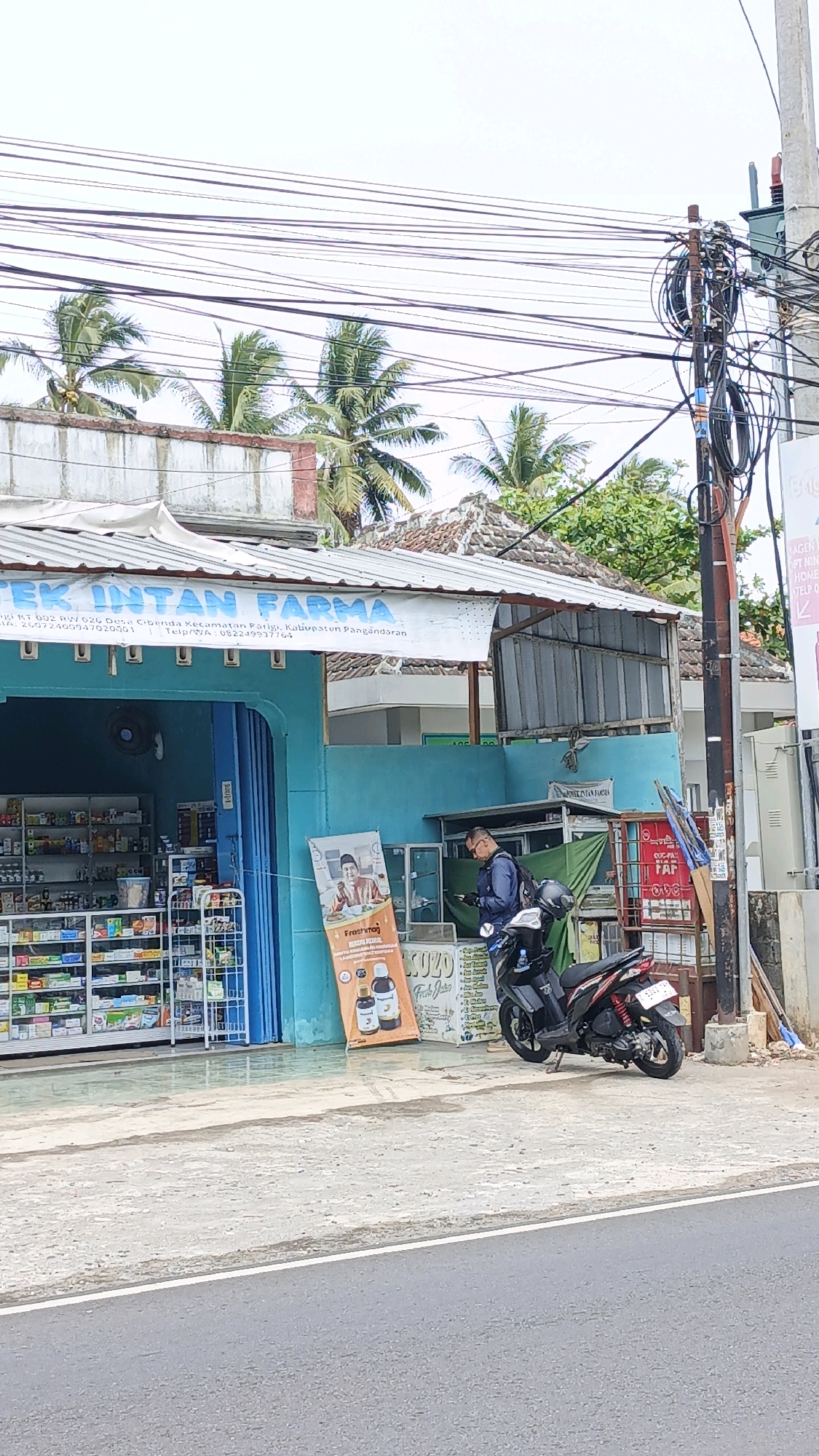 The image depicts an outdoor scene in Cibenda, Indonesia, in front of a pharmacy named "APOTEK INTAN FARMA." The pharmacy has a bright blue facade with a white awning displaying the pharmacy's name and address. Shelves stocked with various products are visible through the open doorway. In front of the pharmacy, a man in a blue jacket is standing near a parked motorcycle, looking at his phone. A banner with an image of a man in traditional attire and text is displayed on an easel to the left of the doorway. A motorcycle is parked on the sidewalk in front of the pharmacy. Coconut trees and power lines are visible in the background, suggesting a tropical or subtropical environment. The overall scene appears to be a typical street view of a commercial establishment in a residential or mixed-use area.