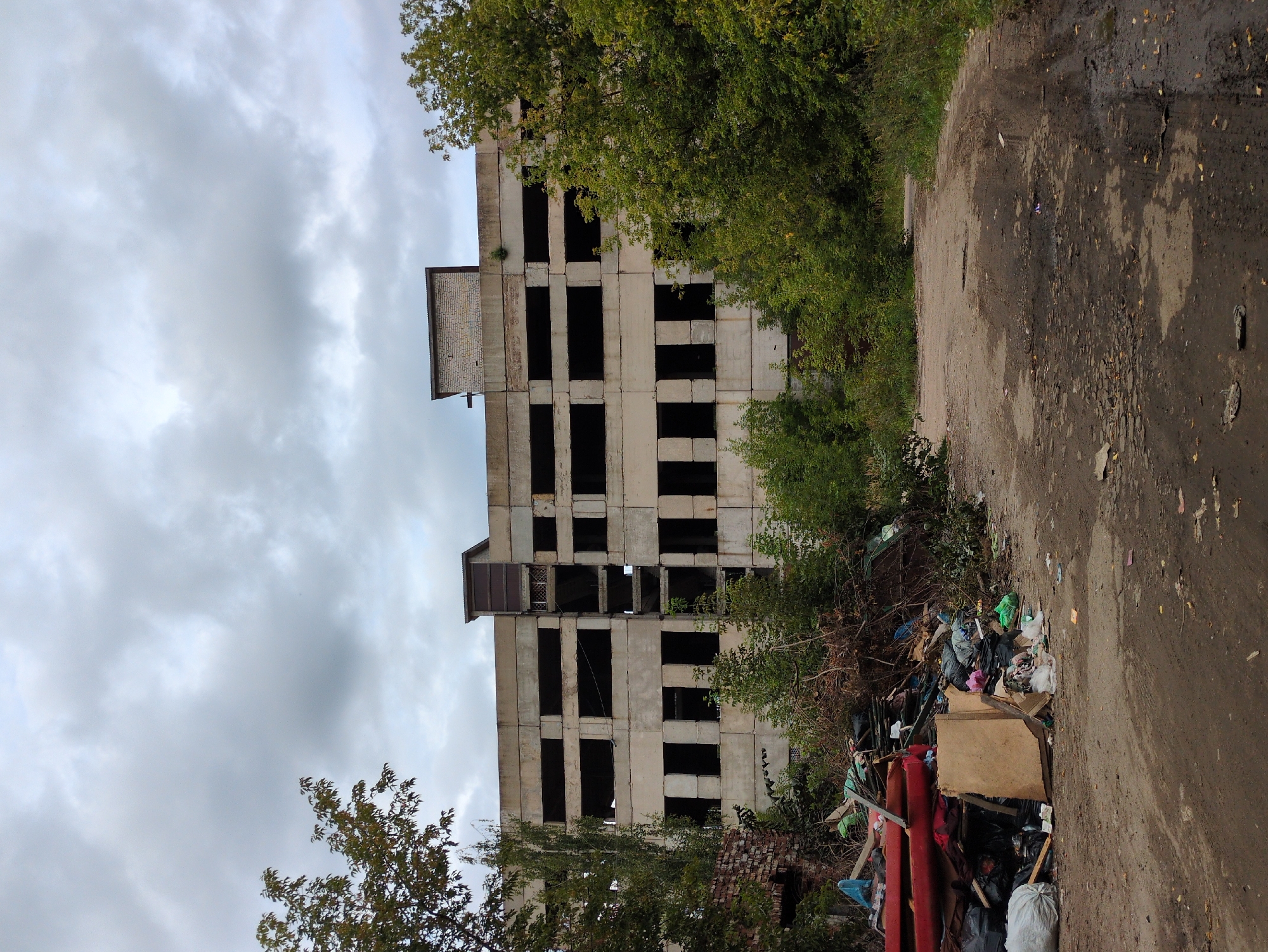 This image shows an abandoned, multi-story concrete building in Kulebaki, Russia. The building has a stark, industrial appearance with numerous dark, empty window openings. In the foreground, there is a large pile of debris, including plastic bags, wood, and other refuse, scattered across a dirt path. Overgrown vegetation surrounds the building, and the sky is overcast with gray clouds. The overall scene conveys a sense of neglect and decay.