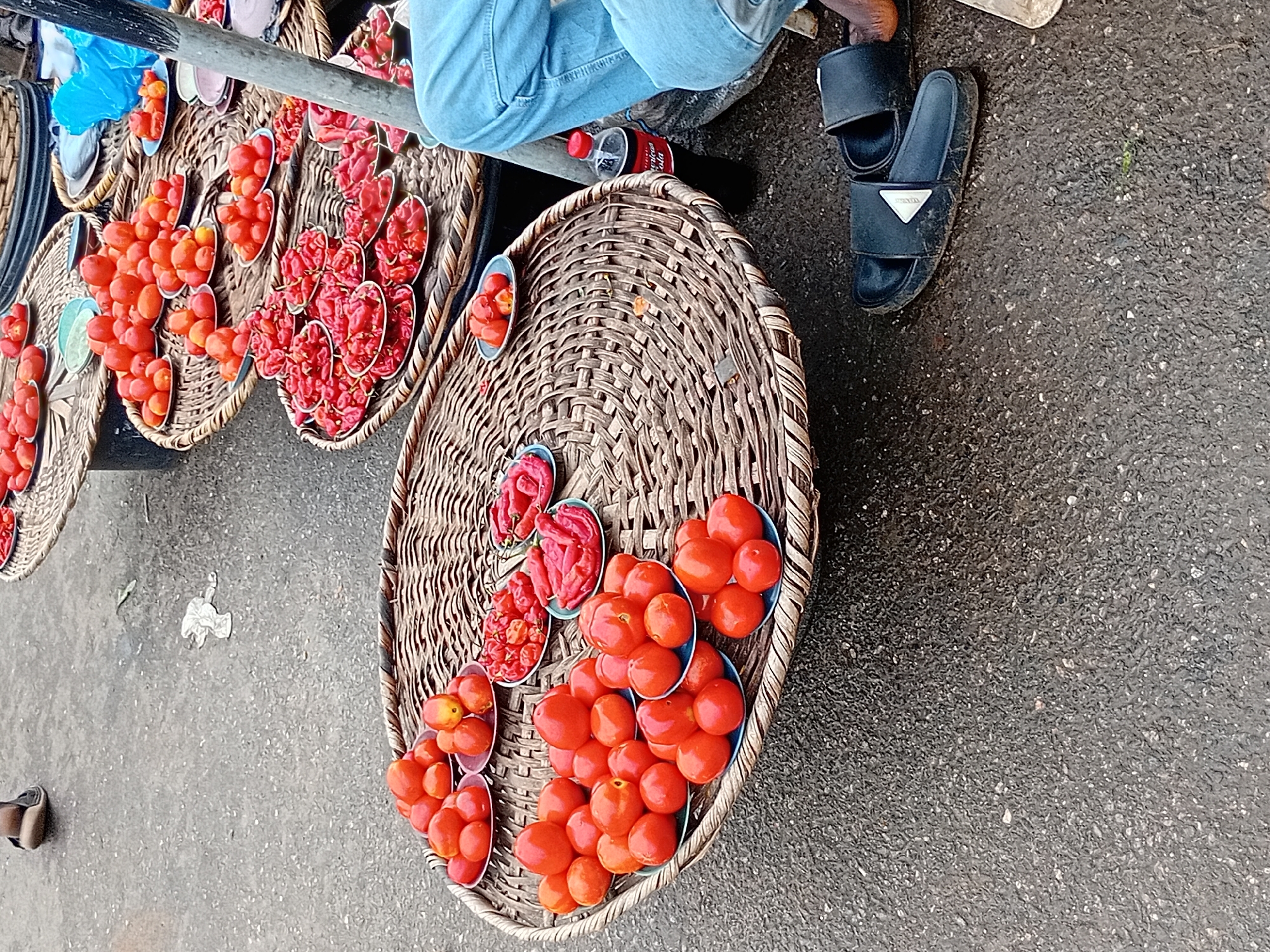 The scene is a street-level shot in Ikorodu, Nigeria. Wicker baskets are displayed on the ground, containing small blue bowls filled with red produce, possibly tomatoes and peppers. A person wearing denim jeans sits near the baskets. Black sandals are visible on the ground. The background is a paved surface, possibly a sidewalk or street. A metal pole or bar is visible near the top of the frame. A bottle of Coca-Cola is situated between the baskets.