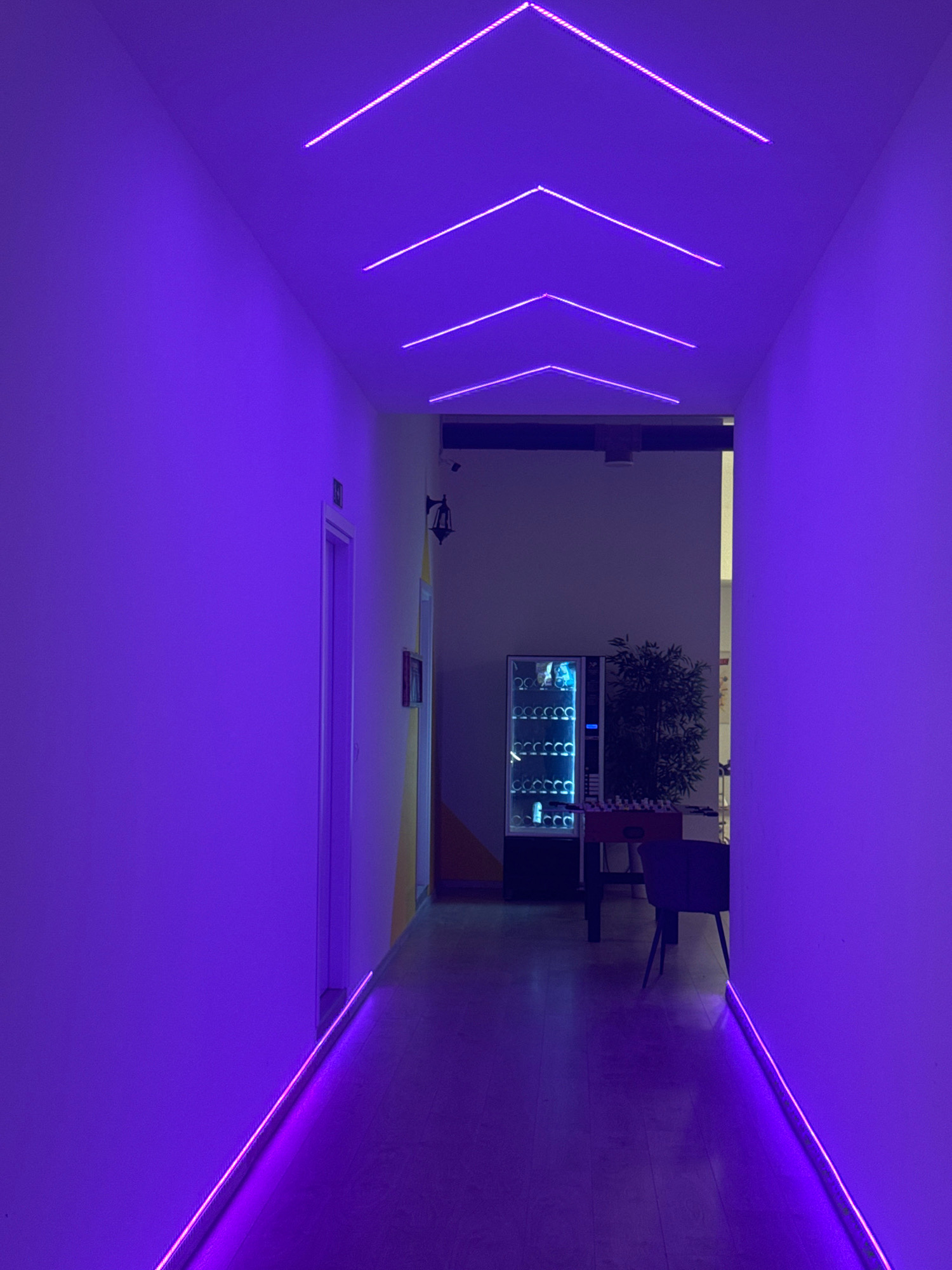 The image depicts a narrow, modern hallway illuminated by vibrant purple LED lights. The lights are installed in a geometric pattern on the ceiling, forming multiple V-shapes that converge towards the back of the hallway. A continuous line of the same purple LED lighting runs along the base of the walls, casting a glow on the wooden floor.
On the right side of the hallway, a white wall transitions to a lighter color towards the back. On the left, a closed door with a white frame is visible. Further down the hallway, on the right, a vending machine is situated, displaying various items through its glass front. Next to the vending machine, a foosball table is set up with a dark-colored chair placed behind it. A potted plant stands to the right of the vending machine. In the background, other doorways and architectural elements are dimly visible.
The overall impression is one of a contemporary and atmospheric space, likely a commercial or entertainment venue. Given the location specified, this hallway could be part of a business, a recreational facility, or a public building within the Germasogeia Municipality, Cyprus. The absence of people suggests the image was taken at a time when the area is not in active use or is unoccupied.