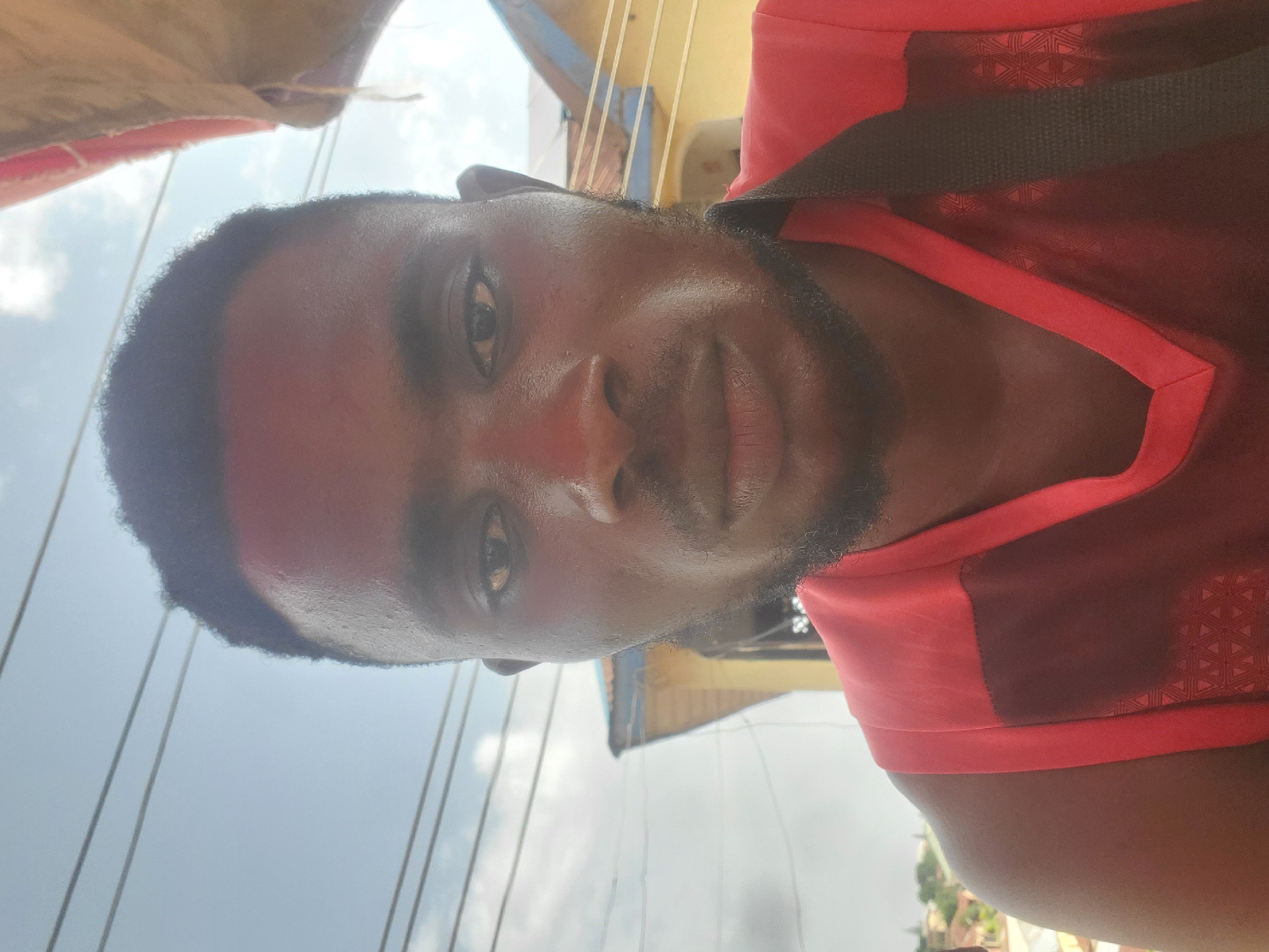 This is a close-up, vertical, selfie-style shot of a dark-skinned young man. He is wearing a bright red t-shirt with dark red accents and a black strap across his chest, possibly from a bag. The camera angle is looking up at him, and the lighting suggests it is daytime, with some overcast sky visible in the background, punctuated by thin, straight lines that could be power lines or wires. The man has short, dark, curly hair, a goatee, and is looking directly at the camera with a neutral expression. His skin appears slightly moist, suggesting he may have been out in the heat. The background is blurred, but hints of yellow and blue structures are visible, along with some greenery. The overall impression is of a casual, outdoor moment captured in Gwong, Nigeria.