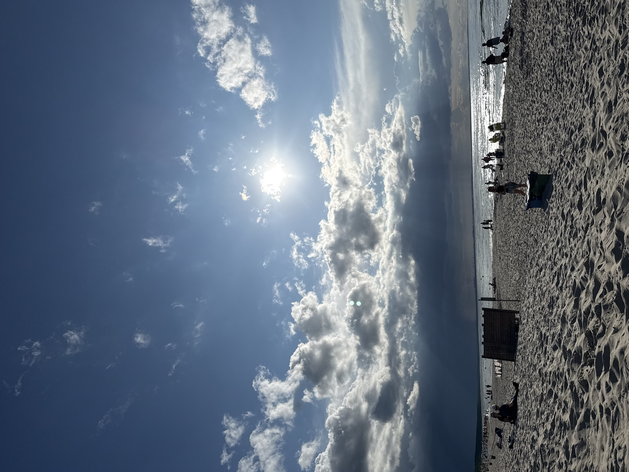 A bright, partly cloudy sky with the sun shining brightly over the Baltic Sea at Międzyzdroje, Poland. The sun is partially obscured by dramatic, dark clouds on the left side of the image, with rays of light breaking through. The sea is calm with gentle waves, reflecting the sunlight. A sandy beach stretches across the foreground, with numerous people scattered across it, some sunbathing, others walking or playing. In the middle ground, a group of people are gathered near the water's edge. Several beach chairs and umbrellas are visible. A wooden structure, possibly a screen or partition, stands on the sand. The overall scene depicts a lively summer day at the beach.