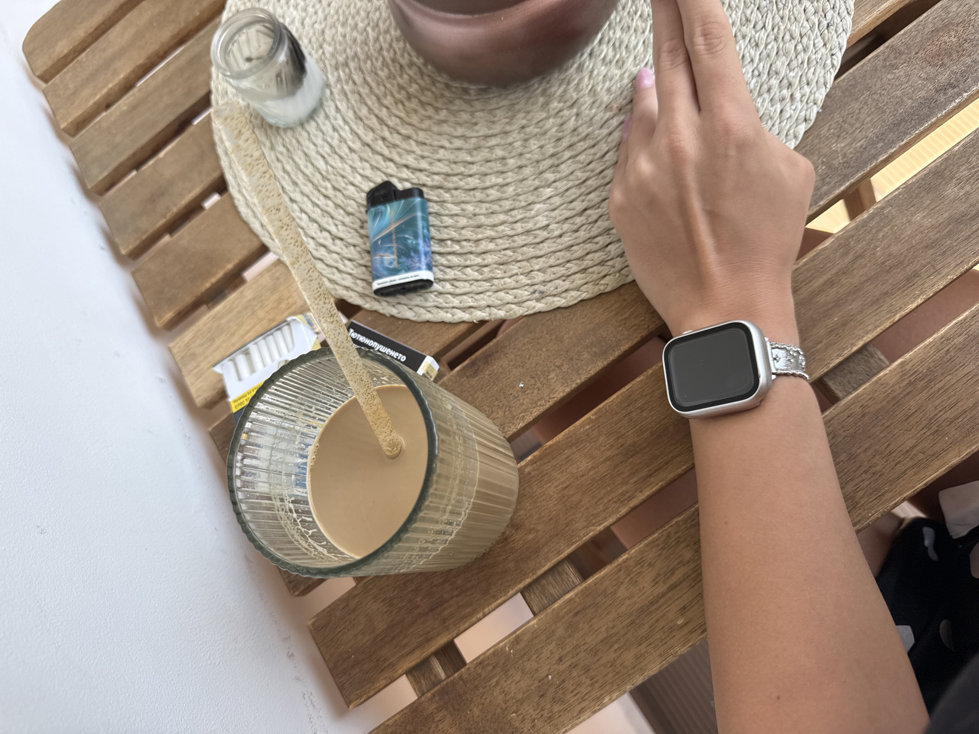 An overhead shot captures a person's arm and hand resting on a wooden slatted table, suggesting a relaxed outdoor setting in Plovdiv, Bulgaria. The scene is brightly lit, indicating daytime.
In the foreground, a ribbed glass contains a light brown, creamy beverage, likely an iced coffee or latte, with a textured straw. Adjacent to the glass, a portion of a cigarette pack is visible, bearing the Bulgarian word "ТЮТЮНОПУШЕНЕТО" (Smoking) along with other smaller, illegible text.
A round, beige woven placemat anchors the center of the frame. On it rests a teal-colored lighter with a swirling, abstract design, and a small, clear glass container, possibly a salt shaker. The person's right hand, adorned with light pink painted fingernails, is positioned on the placemat. A modern silver-toned smartwatch with a dark, square screen and a decorative metal band is worn on the wrist. The rustic wooden table features parallel slats, and a white wall or surface is seen in the background.