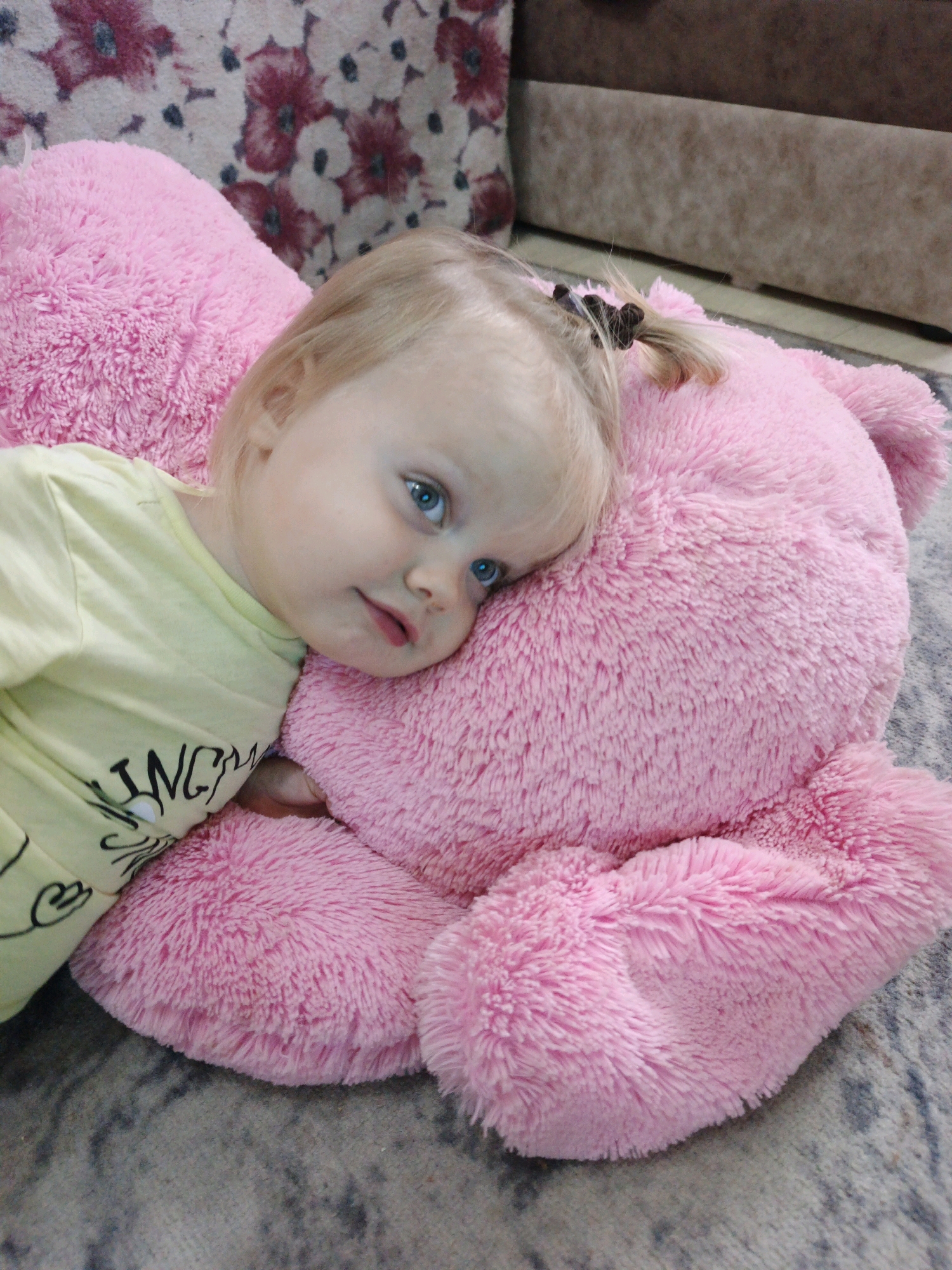 A young, fair-haired child with bright blue eyes rests their head on a large, plush pink teddy bear. The child is wearing a light yellow t-shirt with black lettering. The scene appears to be indoors, with a floral-patterned fabric visible in the background, possibly a sofa or cushion. The child and bear are lying on a textured rug or carpet. The image is a close-up, focusing on the child and the teddy bear. The overall impression is one of comfort and peacefulness. The photograph was taken on a carpeted floor in what is presumed to be a home in Zlatoust, Russia.