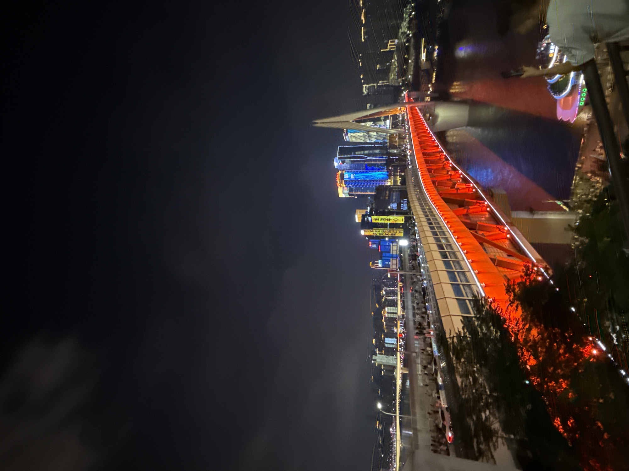 This is a night-time, slightly tilted, wide-angle shot of the Hongyadong Bridge in Yuzhong District, Chongqing, China. The bridge is illuminated with bright red lights, creating a vibrant contrast with the dark sky and the city lights in the background. The bridge's structure is prominently visible, with its cables and supporting beams illuminated. Below the bridge, a river reflects the city's lights. On the bridge itself, there are blurred figures of people walking and cars driving, indicating activity. In the foreground, a person in a light-colored shirt is visible on a balcony, holding up a phone, possibly taking a photo or video of the scene. The backdrop features a skyline of illuminated skyscrapers, with various colors of light, including blue and yellow, emanating from them. The overall atmosphere is one of a bustling urban nightscape.