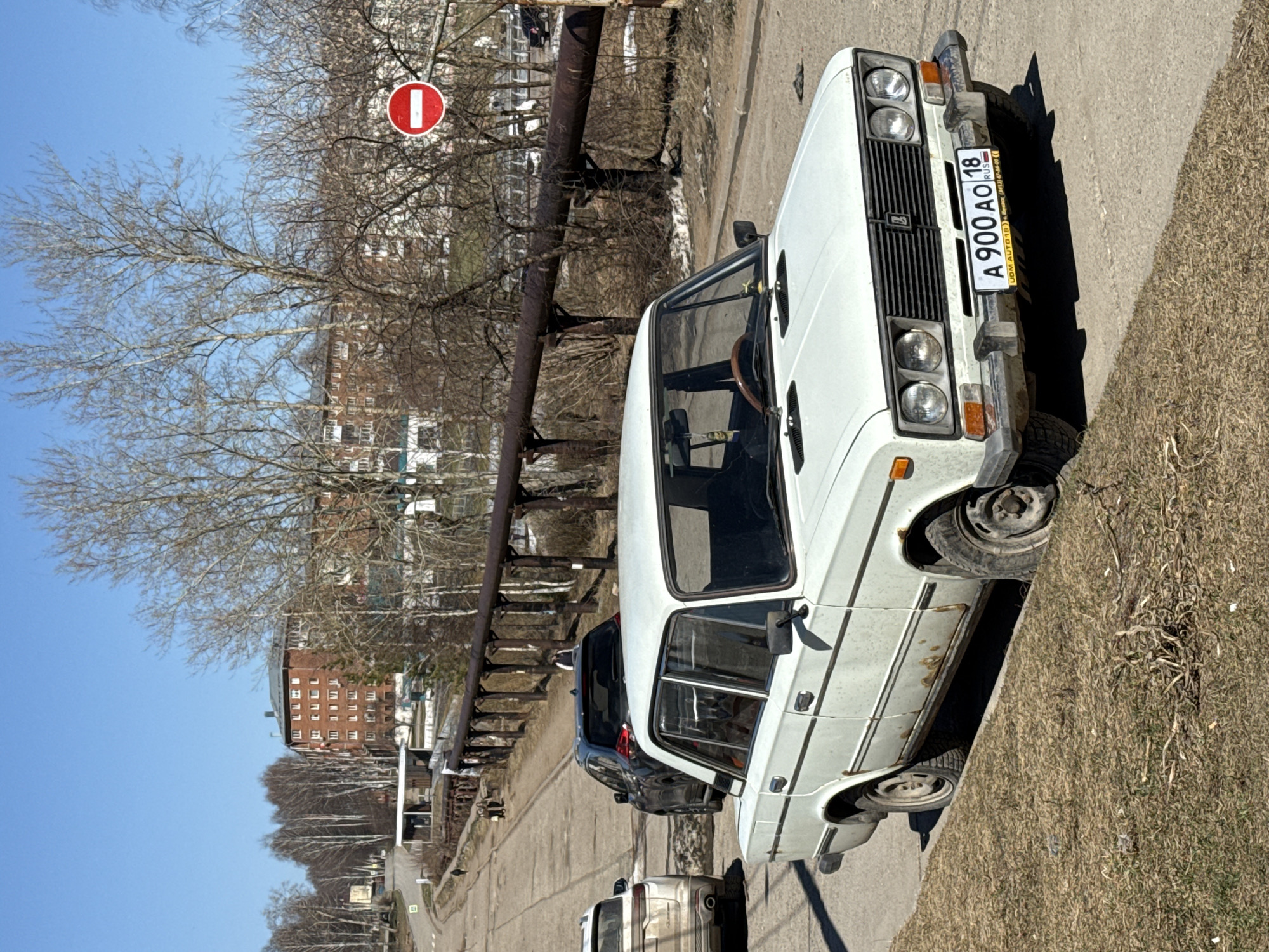 The image shows a white Lada car parked on the side of a road in Votkinsk, Russia. The license plate number is A 900 AO 18. A No Entry sign is visible in the background, along with a brown brick building. Other cars are also parked along the roadside. The environment is an urban area with bare trees. The photograph appears to have been taken during the daytime under clear, sunny skies.