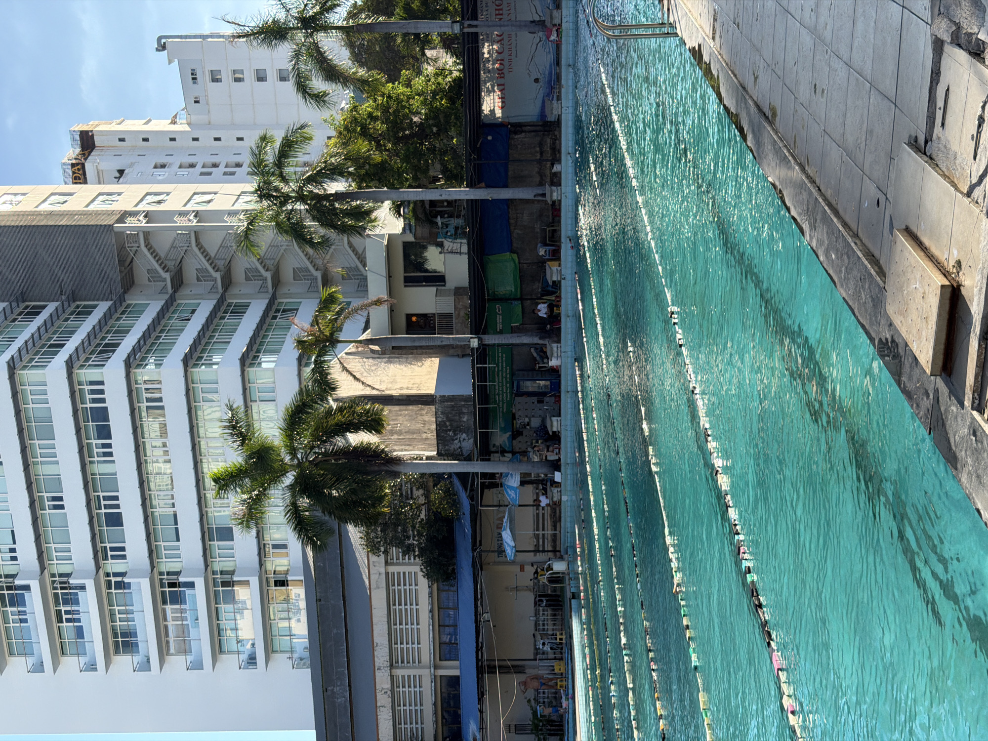 A daytime shot depicts a swimming pool parallel to a building in Nha Trang Ward, Vietnam. The pool is divided into lanes with lane markers, and the water is a light shade of turquoise. Next to the pool is a grey tiled deck.
Adjacent to the pool and behind a row of palm trees, there's a multi-story white building with prominent rows of windows and exterior staircases. To the right of the building, there are other buildings of varying heights and designs, some with visible signs and outdoor seating areas.
People are indistinctly seen in the area between the pool and the buildings. The environment appears to be an urban setting with a mix of commercial and residential structures. The sky is partially visible, suggesting clear weather.