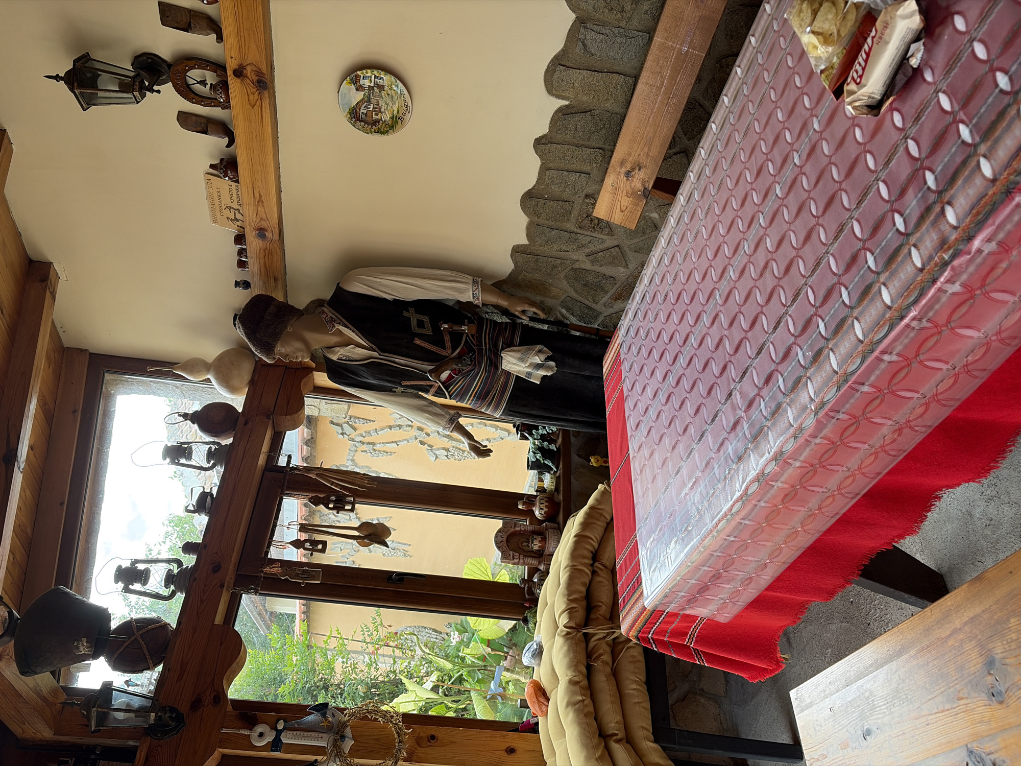 The image shows the interior of a building, likely a restaurant or tourist attraction in Kozlovets, Bulgaria. There's a mannequin dressed in traditional Bulgarian clothing standing near a window. The window offers a view of a garden. A table covered with a red and white patterned tablecloth takes up a significant portion of the frame. On the table are snacks. The room is decorated with various artifacts, including lamps, decorative plates, and wooden carvings, giving it a rustic, cultural feel. The walls are a mix of plaster and exposed stone.