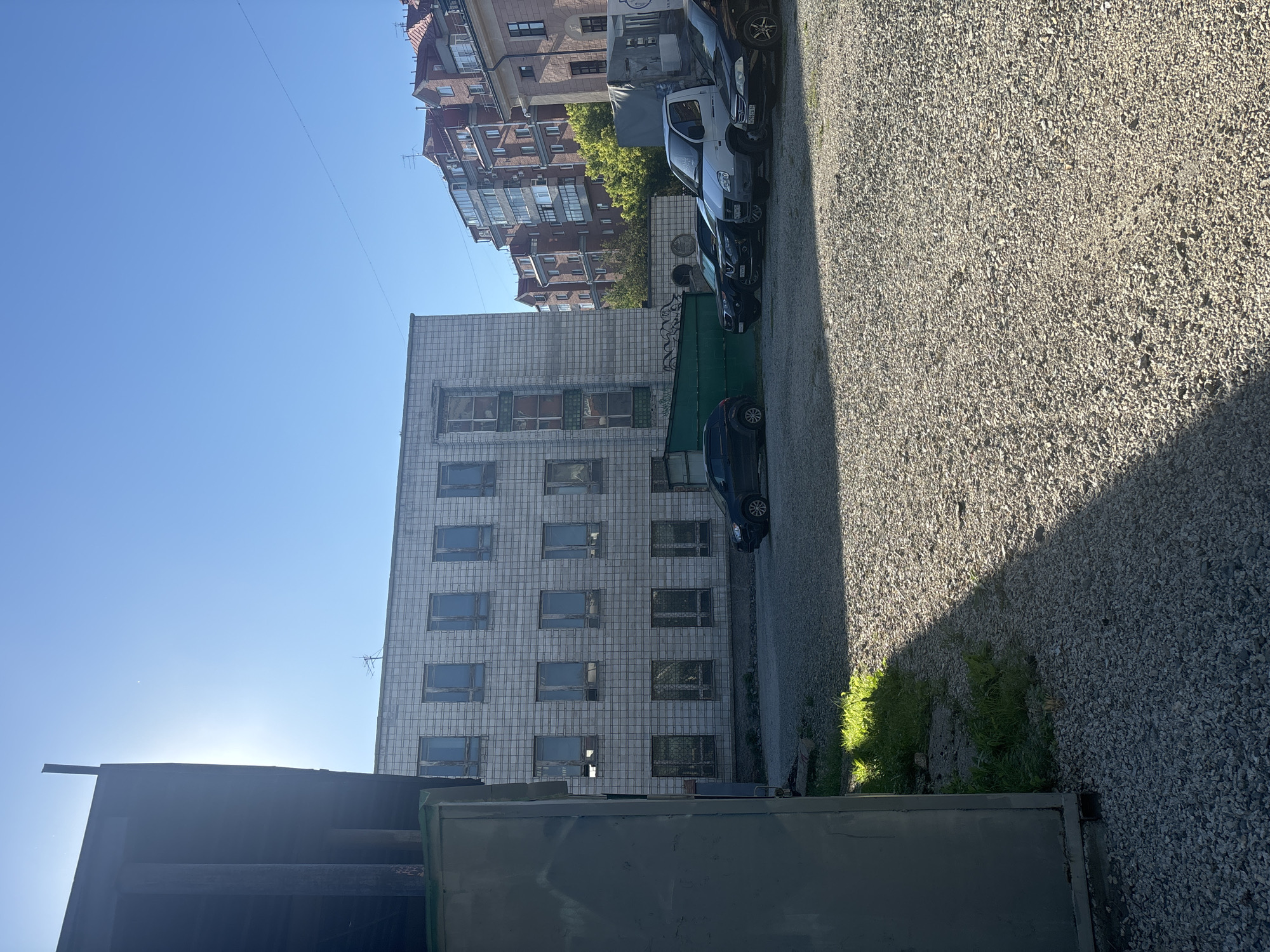 The image shows an outdoor scene in a city environment. In the foreground, a gravel-covered area is visible with several parked vehicles, including a white van, a black SUV, and two other dark-colored cars. To the left, a large, dark, unfinished building structure partially obscures the view. Behind the parked cars, a low, green fence with graffiti is present. Further back, a multi-story building with a light-colored tiled facade stands, featuring numerous windows. Beyond this building, a cluster of taller, reddish-brown residential buildings with multiple windows and balconies can be seen against a clear blue sky. Sunlight is prominent, casting shadows and highlighting the gravel and grass. The scene appears to be a parking lot or an open area adjacent to buildings in an urban setting, likely in Novosibirsk, Russia, given the context provided. There are no people visible in the image.