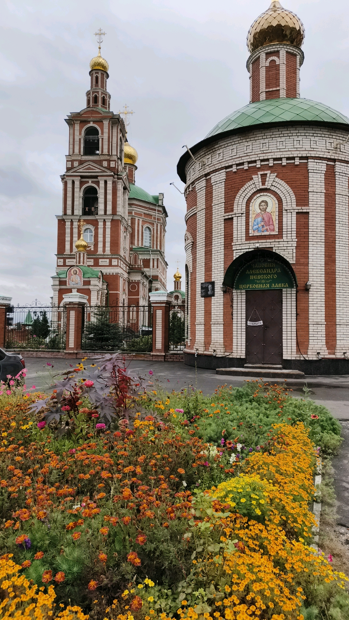 This is a daytime, medium-shot photograph of the Church of St. Alexander Nevsky in Yoshkar-Ola, Russia. The church is made of red and white brick with green roofs and gold domes. A large bell tower is visible on the left, and a round building with an arched entrance is on the right. In front of the church is a colorful flower garden filled with marigolds and other blooming flowers. The sky is overcast, and the overall scene is subdued, with the exception of the bright flowers. A black car is parked on the left side of the image, and a black metal fence runs along the perimeter of the church grounds. There is no visible activity or people in the photograph. The inscription above the arched entrance reads "Church Shop of St. Alexander Nevsky." The words "OPEN" are written on a piece of paper taped to the door.