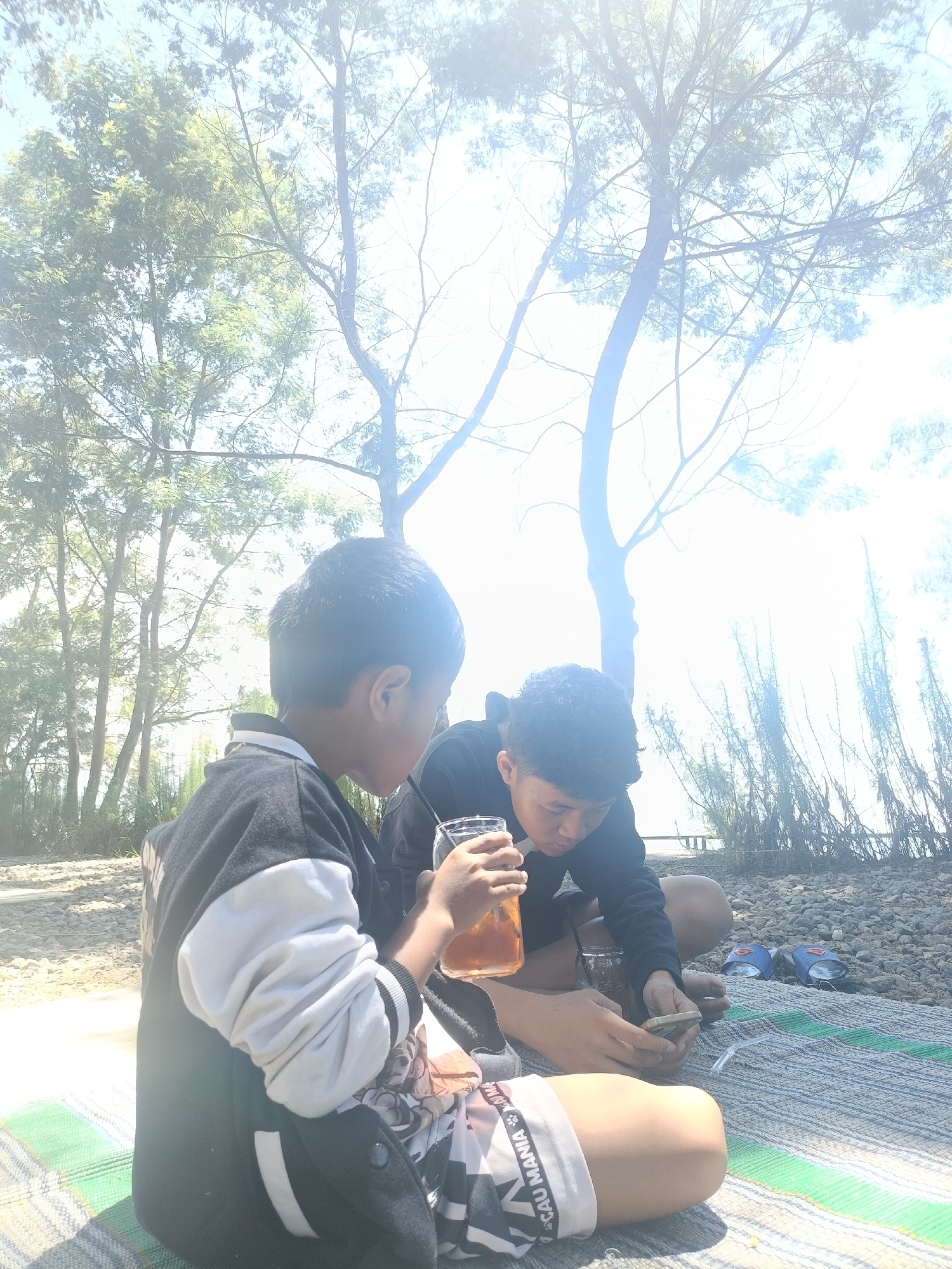 Two young males are sitting on a mat outdoors, in a natural environment. The male on the left, seen from the back, is wearing a black and white jacket and shorts with a pattern. He is holding a glass of iced tea and a straw. The male on the right, facing away from the camera, is wearing a black long-sleeved shirt and shorts. He is holding a mobile phone and a small glass. The background features trees and tall grass, with a body of water visible in the distance. The scene suggests a picnic or leisure activity in a park or by a lake. The location is identified as Kopeng, Indonesia.