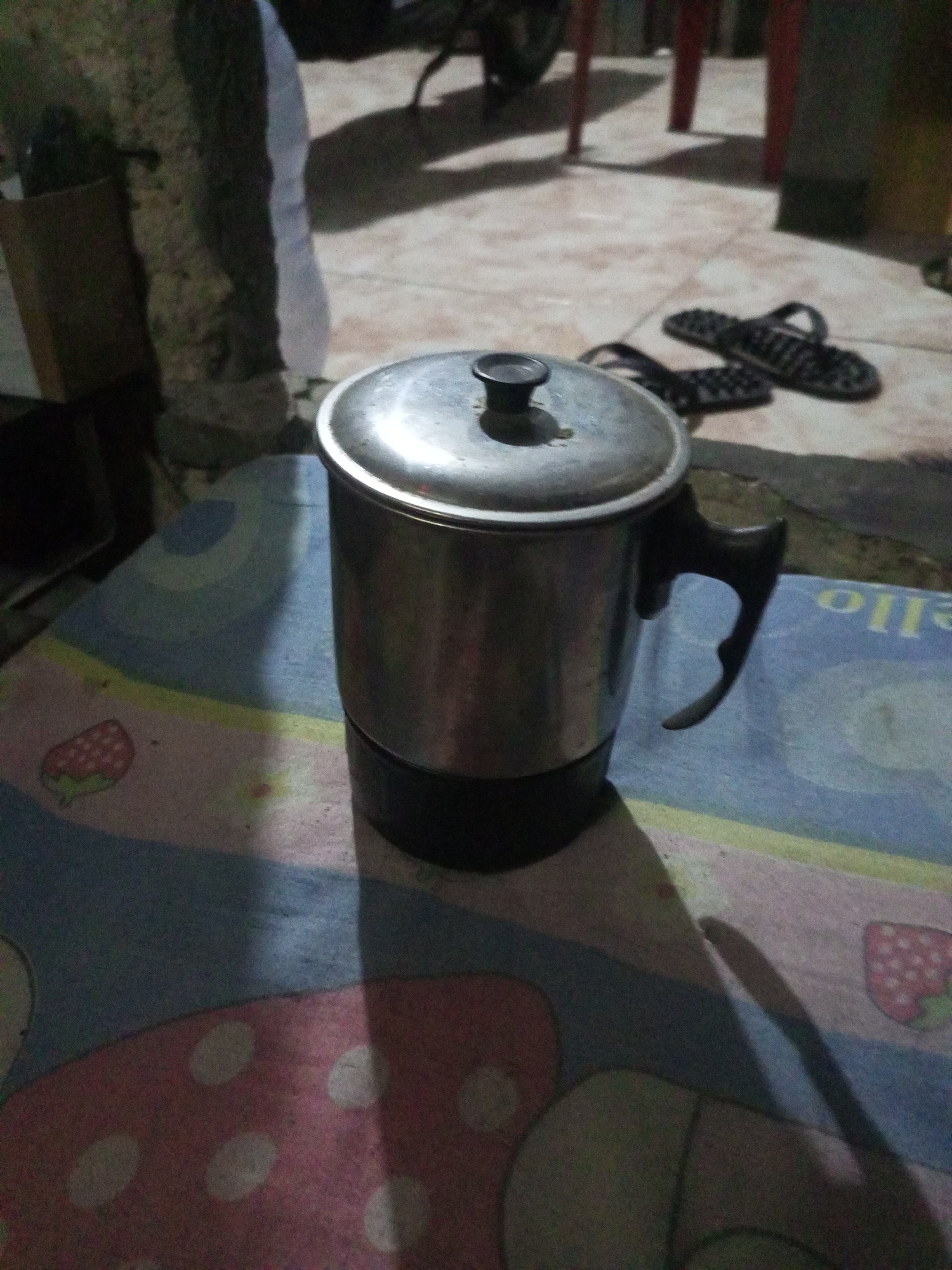 The image shows a stainless steel pot with a lid and handle resting on a colorful mat with a strawberry pattern. In the background, a pair of black sandals are visible on a tiled floor. The scene appears to be outdoors, possibly in a residential area.