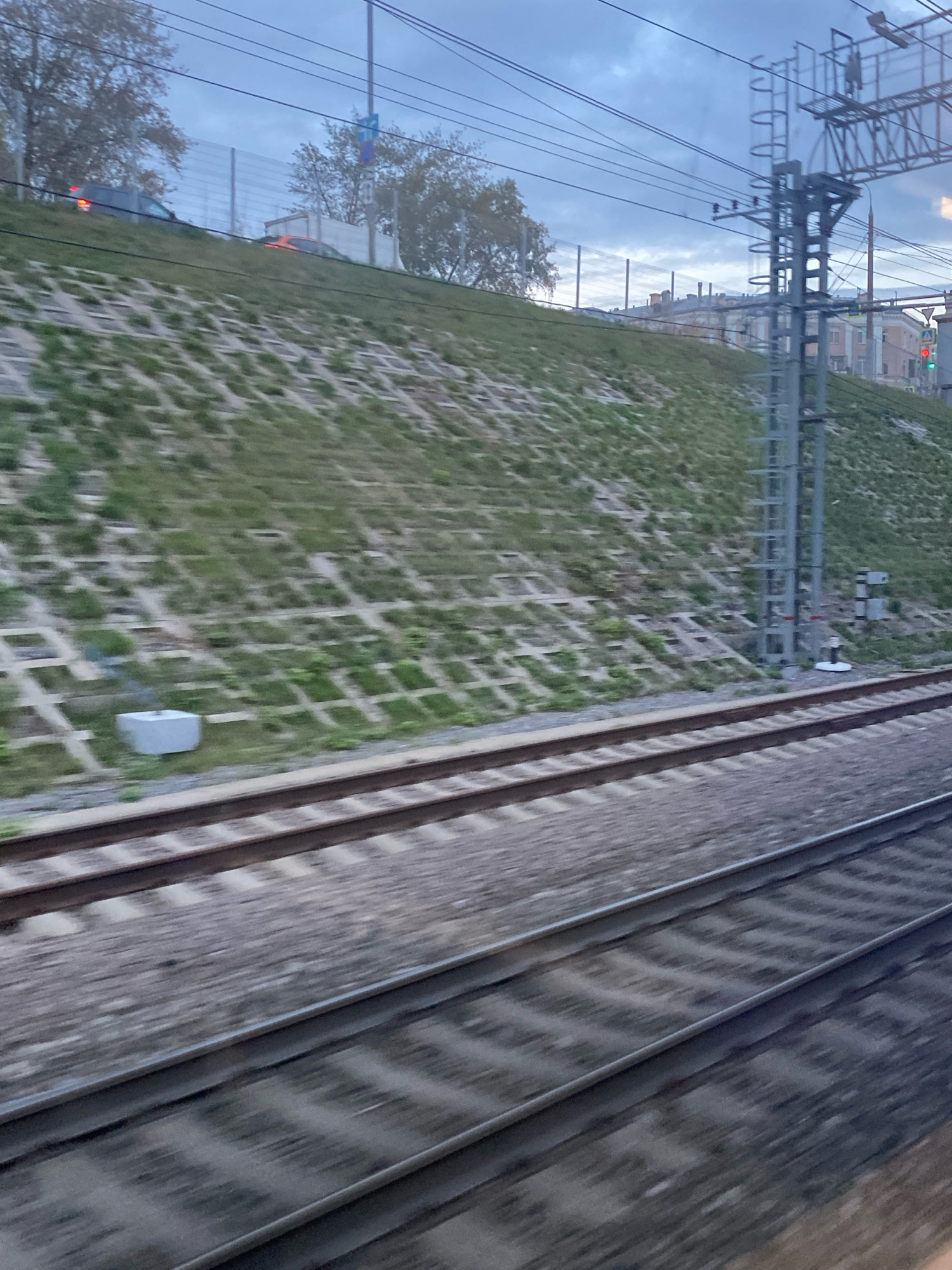The image captures a dynamic scene of railway tracks from the perspective of a moving train, likely in an urban or suburban area of Moscow, Russia. The foreground is dominated by the blurred, parallel lines of the train tracks, indicating motion. Beyond the tracks, a grassy embankment rises, reinforced with a grid of concrete structures designed to prevent erosion. This embankment is partially covered with vegetation.
Further up the embankment, a series of buildings with muted colors and architectural features suggesting an older style are visible. Some of these buildings appear to be multi-story residential or commercial properties. A tall, metallic pylon, likely part of an overhead electrical system for the trains, stands prominently on the right side of the image. There are also traffic lights visible in the distance, with one showing a green light.
In the background, trees with sparse foliage line the top of the embankment, hinting at an autumn or late season setting. The sky is overcast with a mix of gray clouds, creating a soft, diffused light. The overall impression is one of travel through a developing urban landscape, characterized by infrastructure, nature, and human habitation. No people are clearly discernible in the image due to the motion blur and distance.
