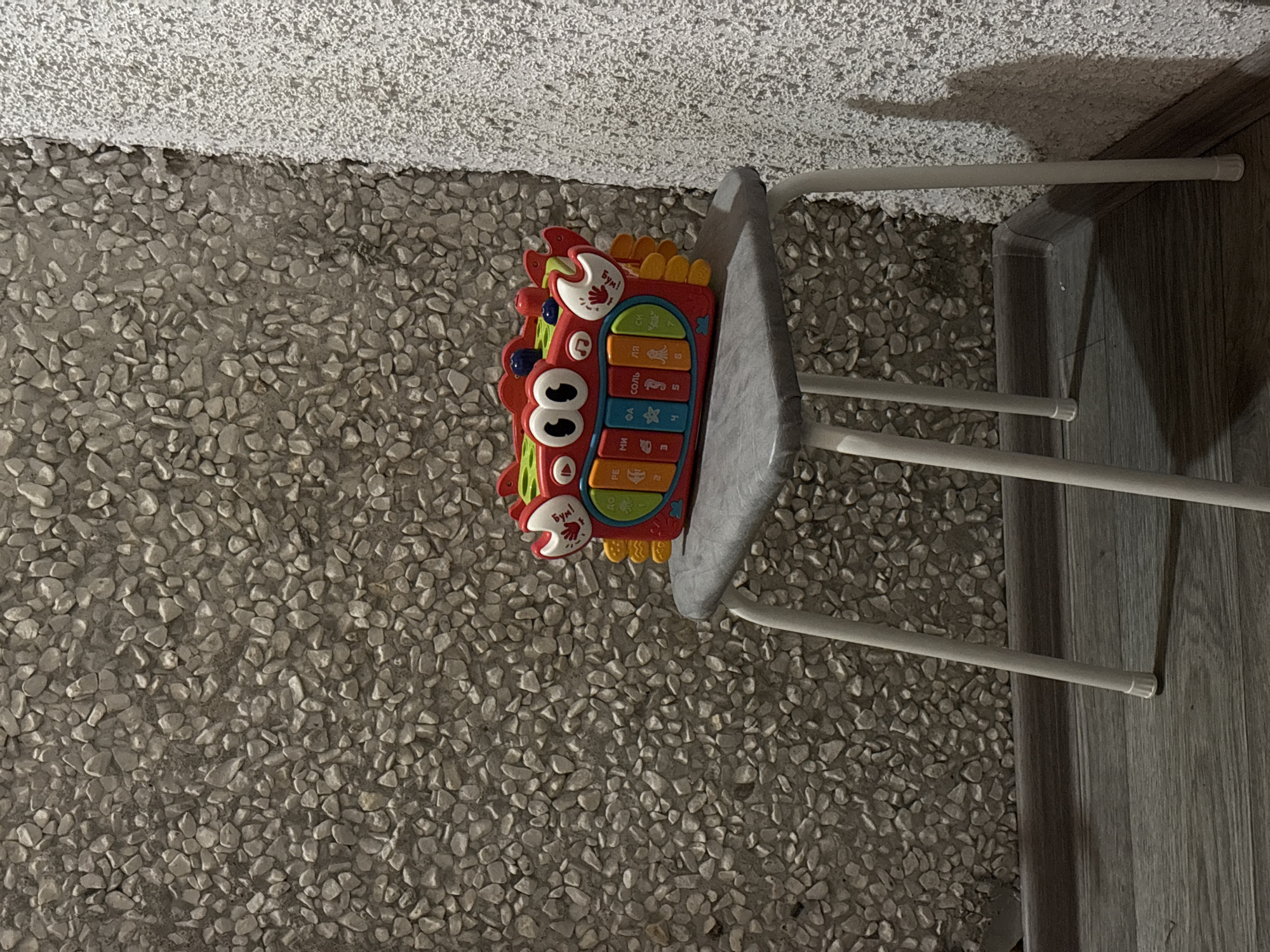 A red, crab-shaped toy keyboard with blue eyes and yellow appendages sits on a gray stool. The stool has thin white legs and is placed on a dark gray wooden floor. The background consists of a textured wall with small, light-colored pebbles and a white, rough-textured wall on the right. The lighting suggests an indoor environment, possibly a room in Астроміцкі сельскі Савет, Belarus.