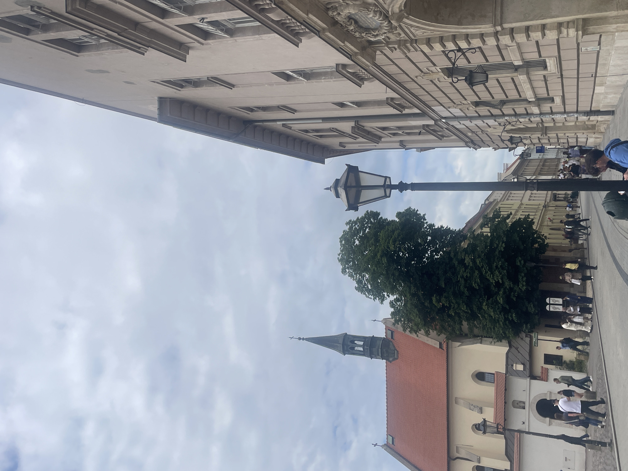 The image depicts a street scene in Krakow, Poland, likely in the historic Old Town. On the left, a building with a red tiled roof and a pointed steeple, possibly a church, is visible. A large green tree stands to the right of this building. A lamppost with a traditional design is prominent in the foreground. To the right, a light-colored building with classical architectural elements, including arched entrances and decorative facades, lines the street. Several people are walking on the cobblestone street and sidewalk, some in groups, and others individually. The sky is overcast with clouds.