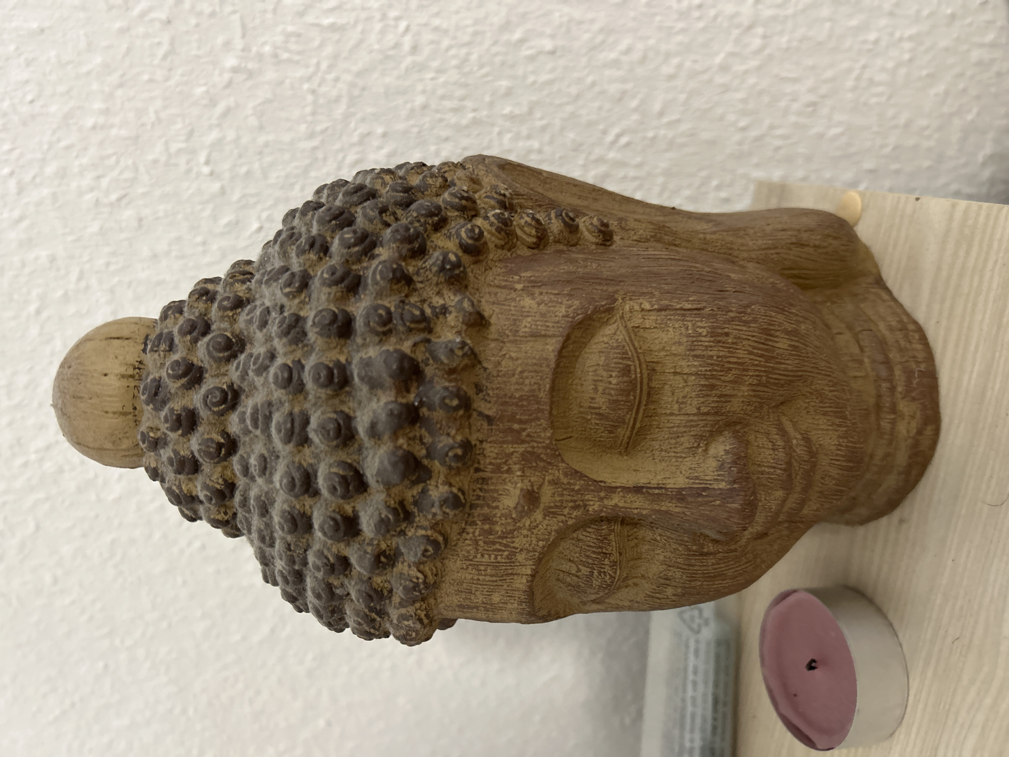 A Buddha head statue stands prominently in the image, positioned on a light-colored, horizontal surface, likely a shelf, which exhibits subtle wood grain. The statue is crafted from a coarse, earthy-toned material, possibly ceramic or composite, displaying a visible vertical striation texture across its face and neck. Its features include tightly coiled, dark brown hair or bumps covering the scalp, and a prominent ushnisha (cranial protuberance) topped by a distinct, lighter, conical element. The eyes of the statue are closed, conveying a serene expression, and the facial contours suggest a meditative state. A fine layer of dust or particulate matter is visible on the statue, particularly on the textured hair curls.
To the left of the statue, a small, circular tea light candle in a silver-colored metal cup rests on the same surface. The candle wax is purple, and its black wick, centrally located, appears extinguished. Behind the candle, partially obscured and out of focus, are faint outlines of what appear to be rectangular objects. The background is composed of a uniformly white wall with a distinct, uneven textured finish, resembling stippled or roughcast plaster. A small, pale yellow, irregular mark or blob is visible on the shelf surface to the lower right of the statue's base. No people or discernible actions are present in the scene, which appears to be an indoor setting.