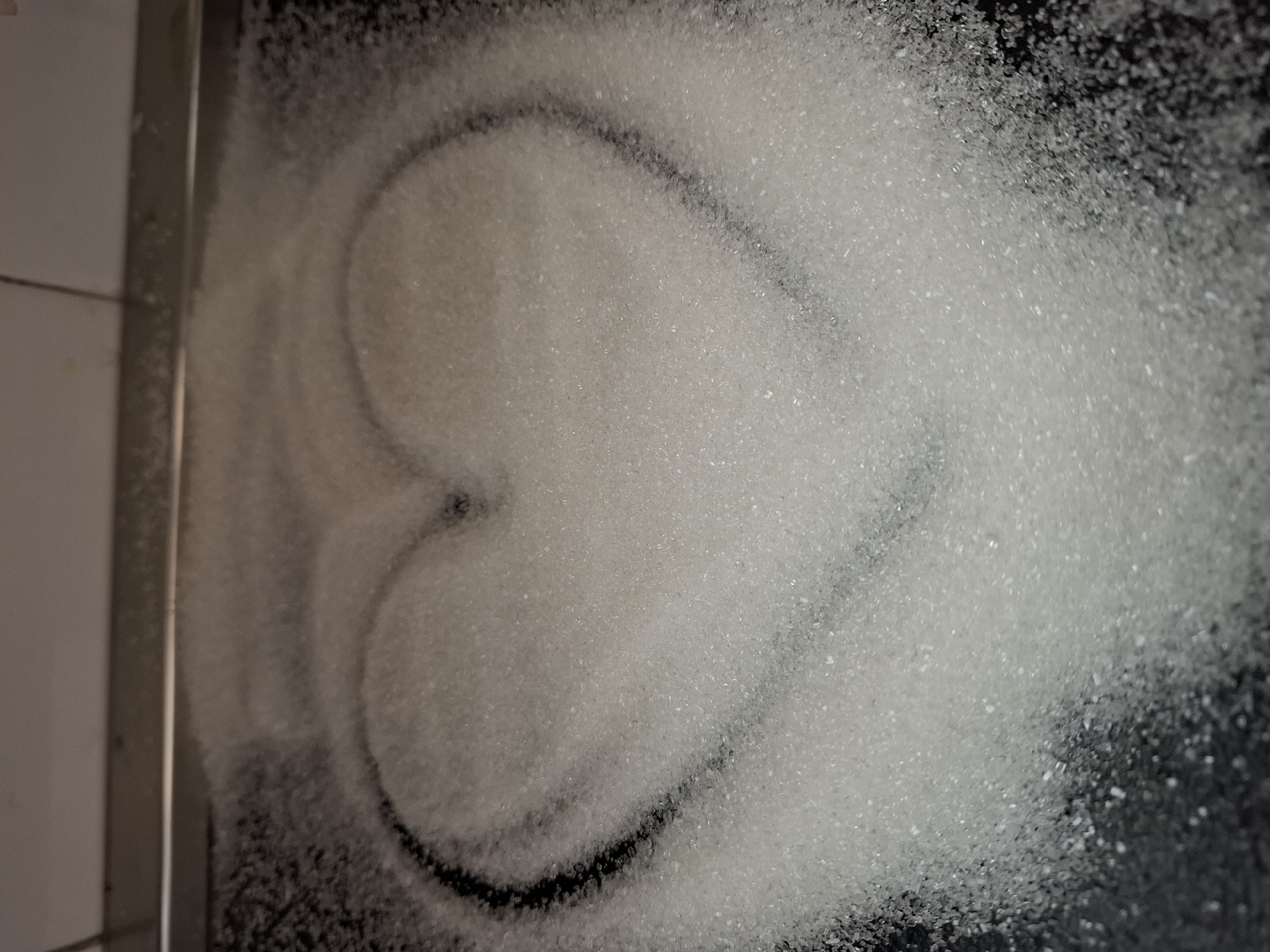A close-up, slightly angled shot reveals a pile of granulated white sugar on a dark, possibly black, surface. The sugar has been spread out, and a heart shape is faintly outlined within the sugar pile, revealing the dark surface beneath. To the left of the frame, a metallic edge or frame is visible, along with a white, possibly tiled, surface. There are no people visible in the image, nor indications of location details like Novi Sad, Serbia. The primary subjects are sugar and the implied heart shape. The interaction is the shaping of the sugar. The environment appears to be a kitchen or food preparation area.