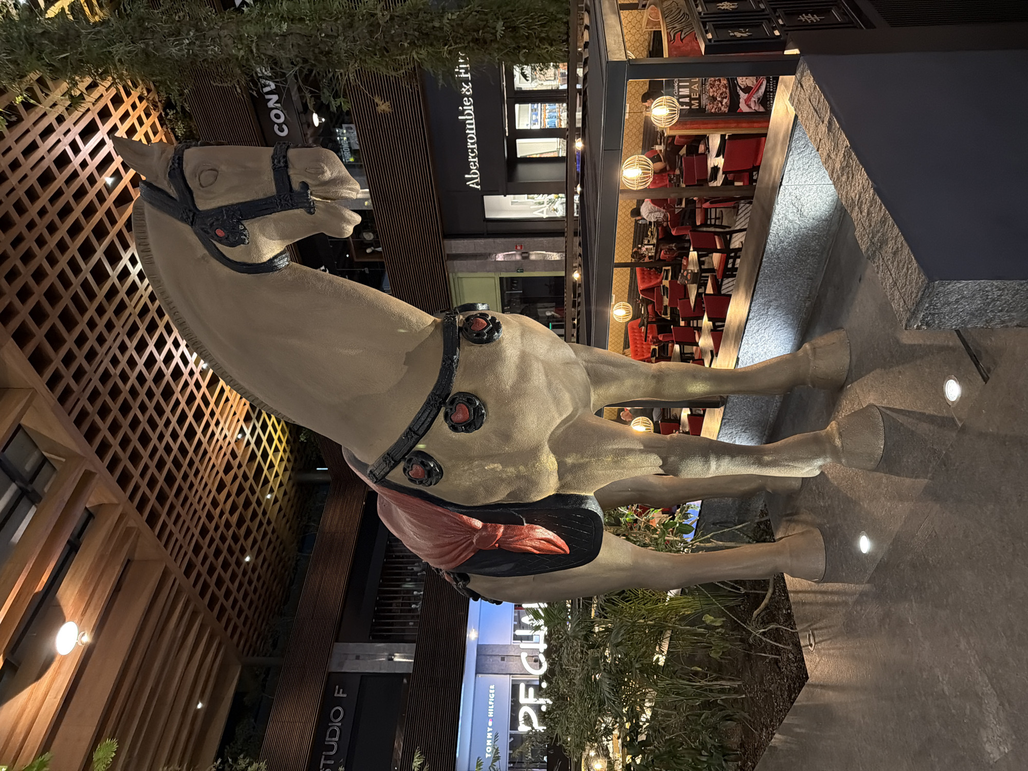 A large, beige statue of a horse stands on a stone platform in an outdoor urban setting. The horse is saddled with a black harness and a distinctive red bow on the saddle. In the background, recognizable retail stores such as "Abercrombie & Fitch" and "Tommy Hilfiger" are visible, as well as restaurants with patrons seated at red chairs. The architecture includes modern buildings with wooden facades and glass windows. The setting is likely the city of Puebla, Mexico.