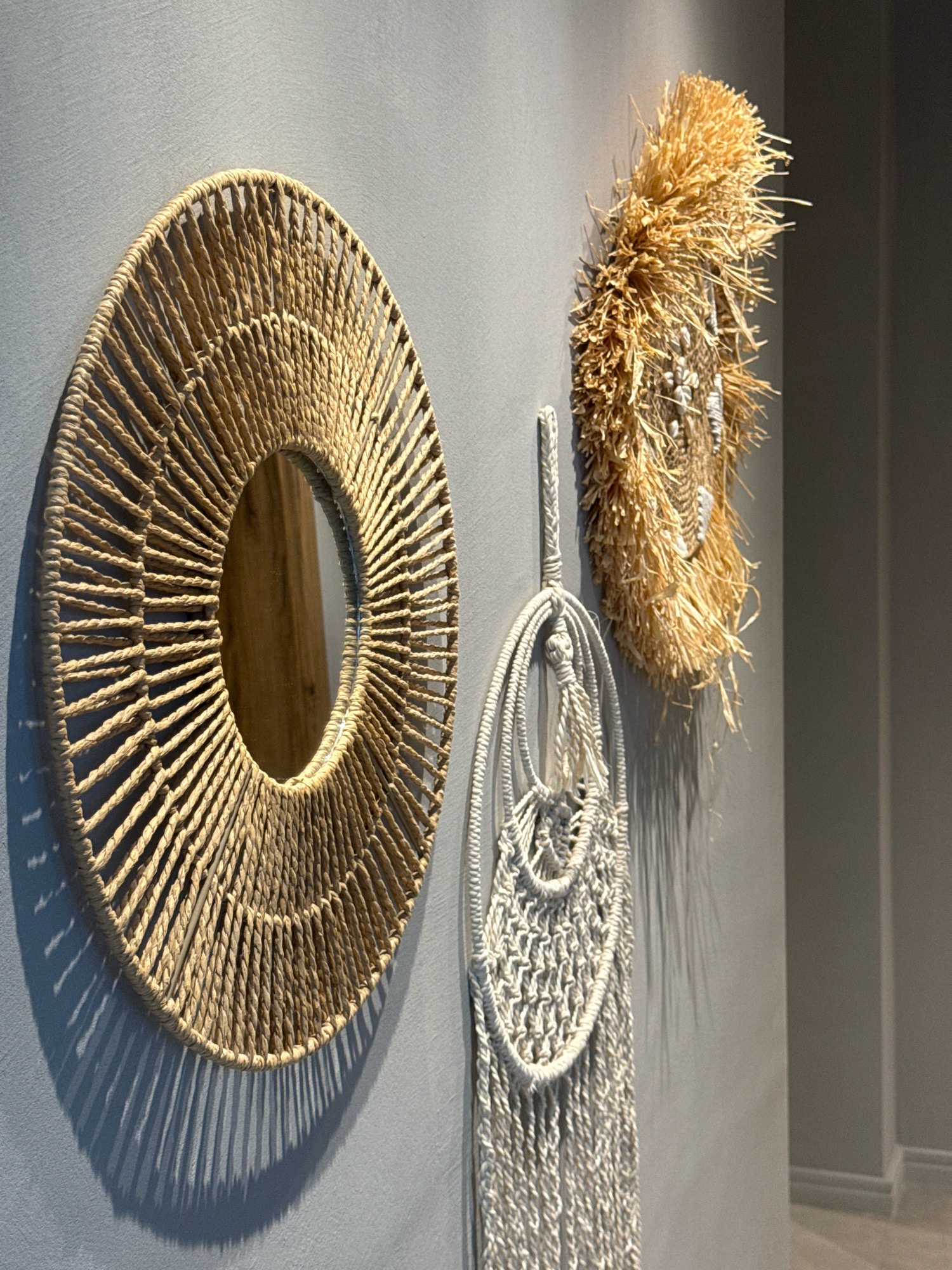 Three wall hangings are displayed on a plain gray wall in what appears to be an indoor setting. On the left is a round mirror framed with tightly woven natural fibers, resembling rope or straw. The weaving extends radially from the mirror's center, creating a sunburst effect. To the right of the mirror is a smaller, more textured circular object made of dried straw or reeds, with some white beads or small shells incorporated into its design. Hanging below this straw piece is a long, white macrame tapestry featuring intricate knotwork and fringed ends. The lighting casts distinct shadows from the objects onto the wall, suggesting a light source from the upper left. The overall aesthetic is bohemian or coastal. There are no people or animals present. The location is not specified in the image, and it is not possible to determine if it is in Sozopol, Bulgaria.