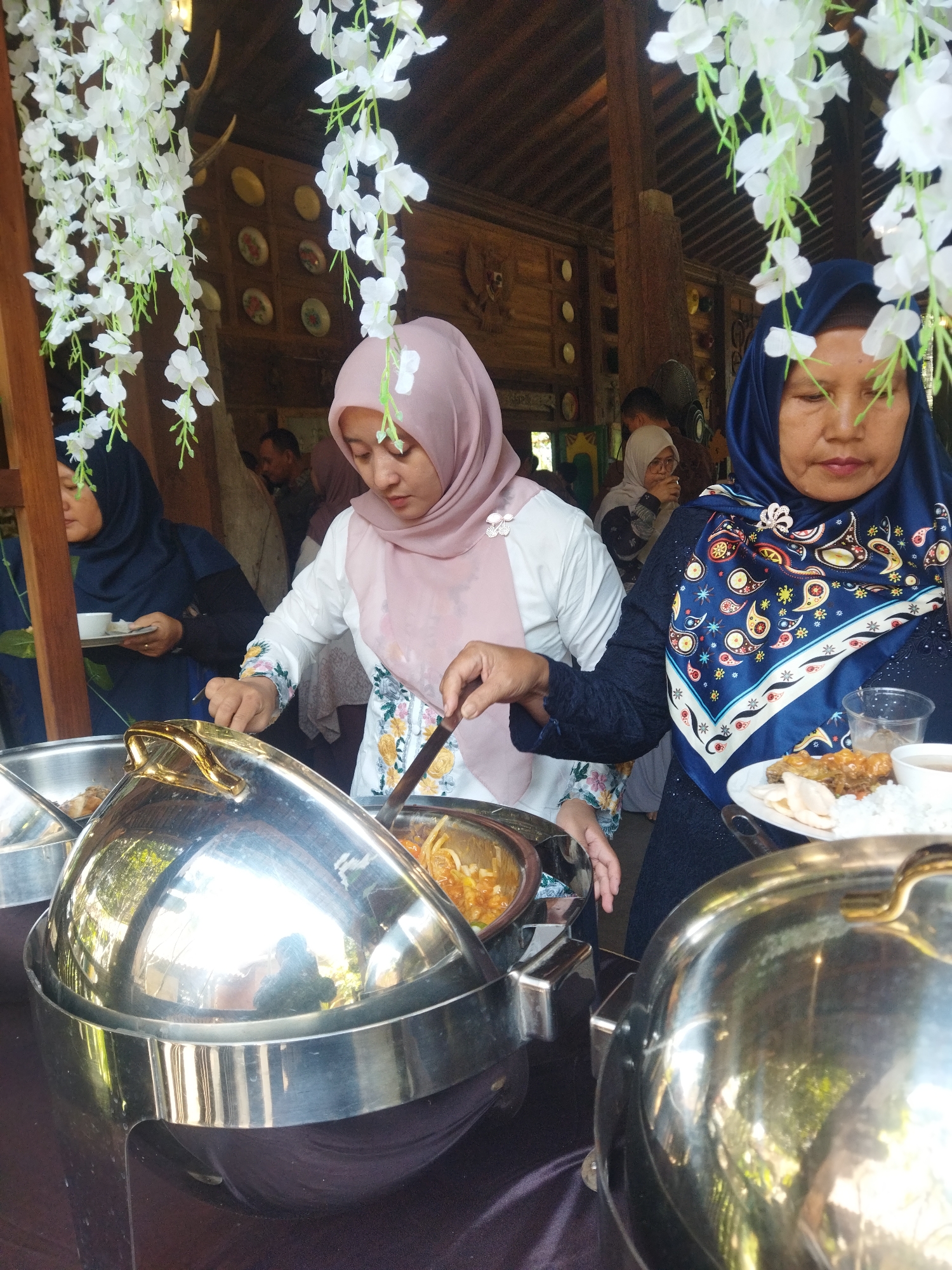 A group of people are gathered around a buffet table at an outdoor event. Two women in headscarves are prominently featured. The woman on the left wears a pink headscarf and a white shirt with floral embroidery, and is using a serving spoon to dish out food. The woman on the right wears a blue headscarf and a dark blue dress with a patterned scarf, and is holding a plate of food. The buffet table has large silver chafing dishes, one of which is open, revealing food. White floral decorations hang from above, suggesting a festive or ceremonial occasion. The background shows wooden structures and a glimpse of greenery, hinting at an outdoor setting in a tropical or subtropical region. The presence of decorative antlers hanging from the ceiling suggests a possible cultural theme or a specific type of venue. The overall scene depicts a food service setting at an event.