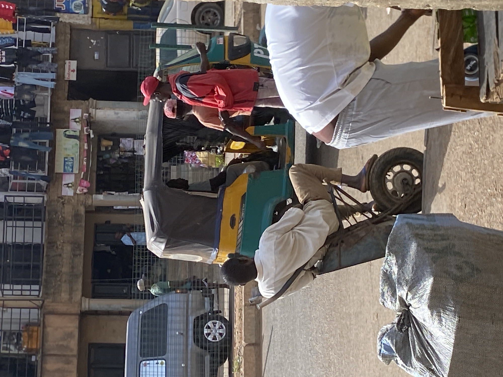 A daytime street scene in Bukuru, Nigeria features a three-wheeled vehicle (tuk-tuk) carrying passengers. A man in a red cap and red jacket is a passenger, and another man in an orange shirt is behind him. The driver is wearing a white shirt and light-colored pants. He is sitting on the vehicle, apparently stationary. A man in white clothing is standing next to the vehicle and interacting with the passenger. A bag is near him on the ground. In the background, there are buildings, a parked minivan, and a clothes rack with clothes hanging.