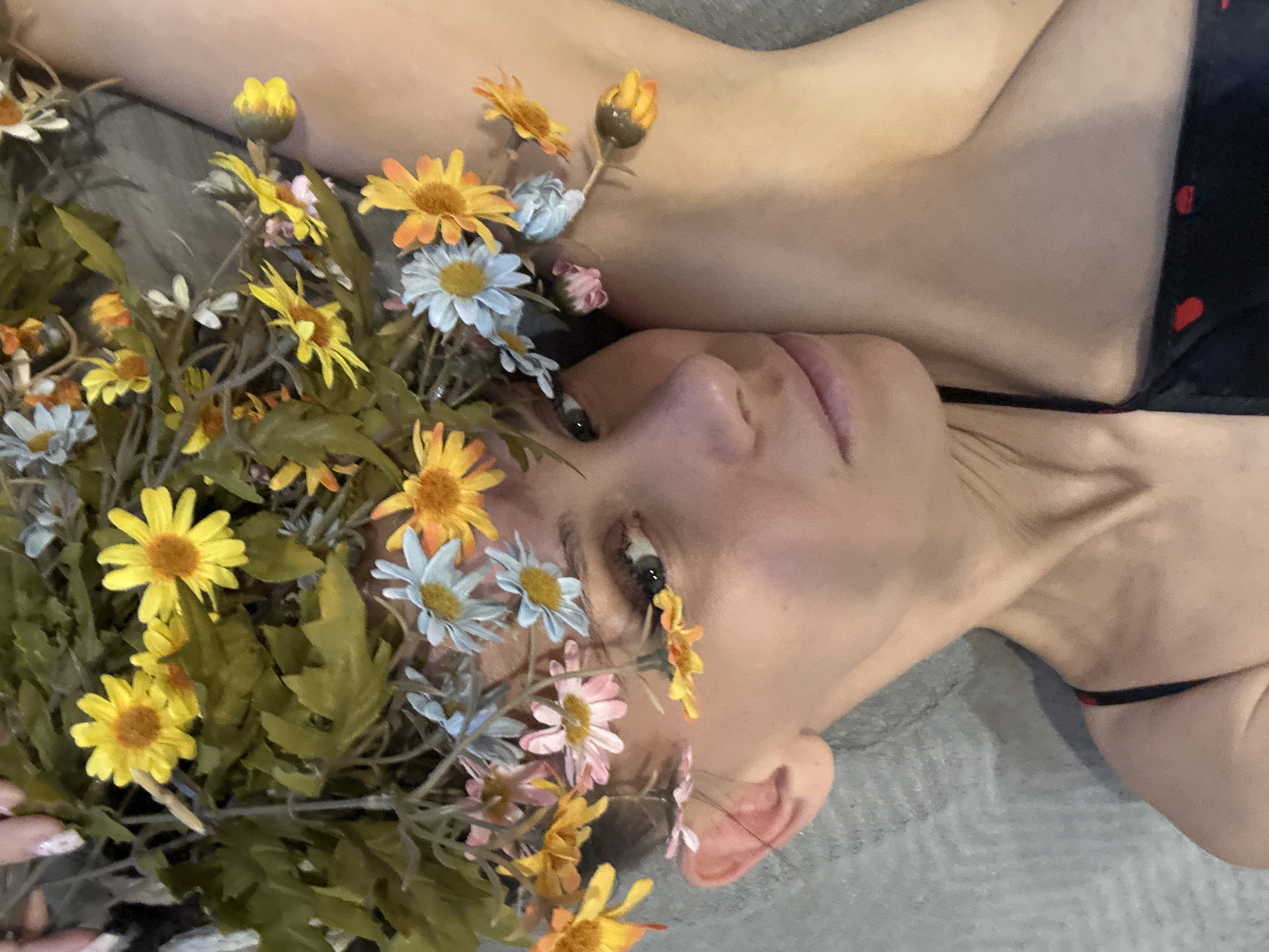 A woman lies on a gray surface, likely fabric, angled towards the viewer, with her head adorned with a profusion of yellow, orange, blue, and pink daisies and green foliage. She is wearing a black garment with red heart details. The woman's face is visible, revealing her features, and her ear is also visible. The context suggests a staged or styled photograph rather than a candid one. There are no indications in the image of Yaroslavl', Russia.