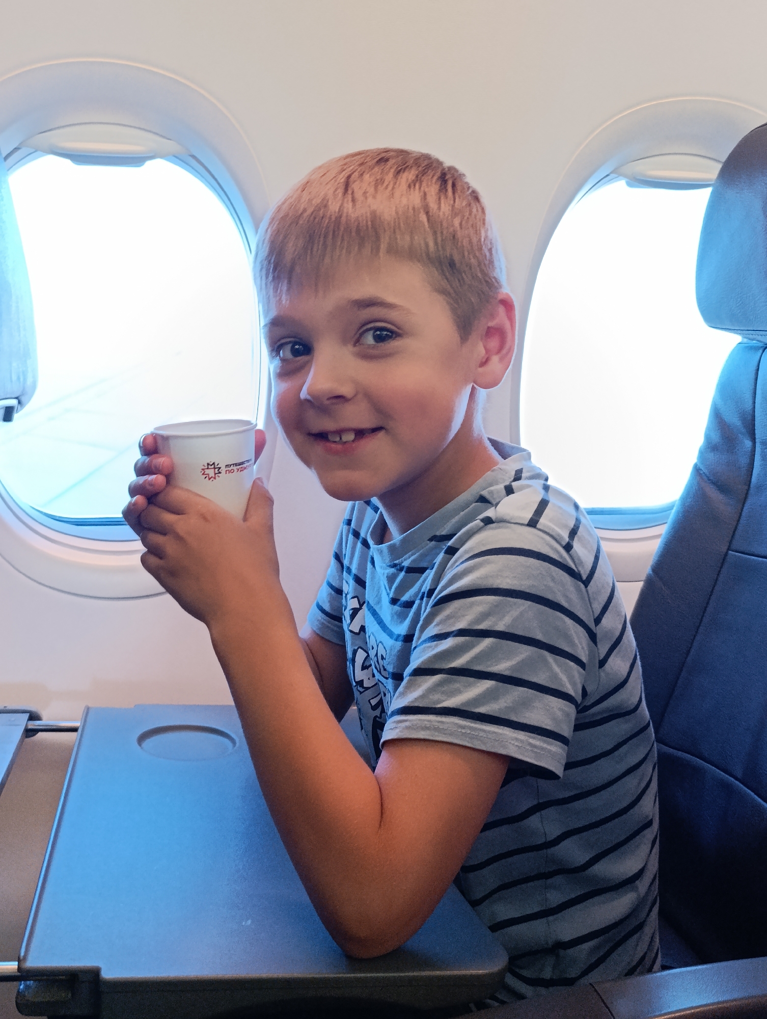 A young boy with short, light brown hair and blue eyes is sitting by an airplane window, holding a white paper cup with red and blue text and a logo. He is smiling and looking at the camera. He is wearing a light blue t-shirt with dark blue horizontal stripes. The airplane's tray table is down in front of him, and an airplane seat is visible to his right. The scene is lit by the bright sunlight coming through the window. This image is not located in Staroe Mart'yanovo, Russia.