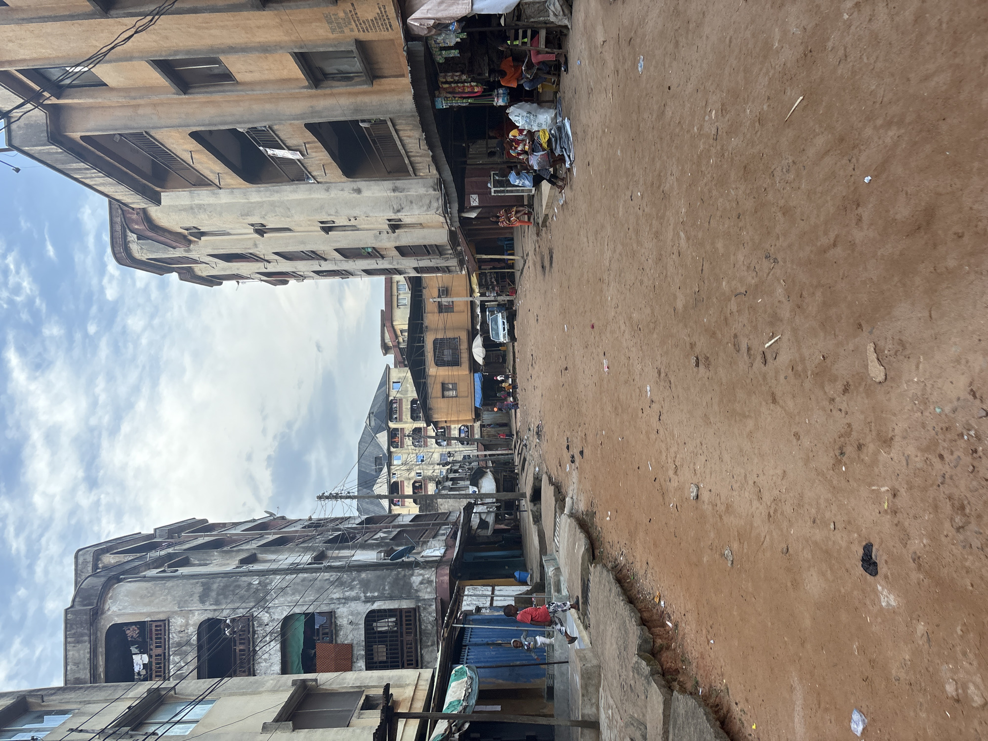 A street scene in Onitsha, Nigeria, features a dirt road lined with multi-story buildings. The buildings are aged, with weathered facades, some showing signs of disrepair. Clothes are visible hanging from balconies and windows. Several small shops or stalls are set up along the sides of the street, with goods displayed outside. People are present, including children walking and adults gathered around the shops. A white van is parked on the street in the distance. The ground is primarily reddish-brown dirt, with some concrete slabs and drainage channels. Debris and litter are scattered across the road. The sky is overcast with clouds.