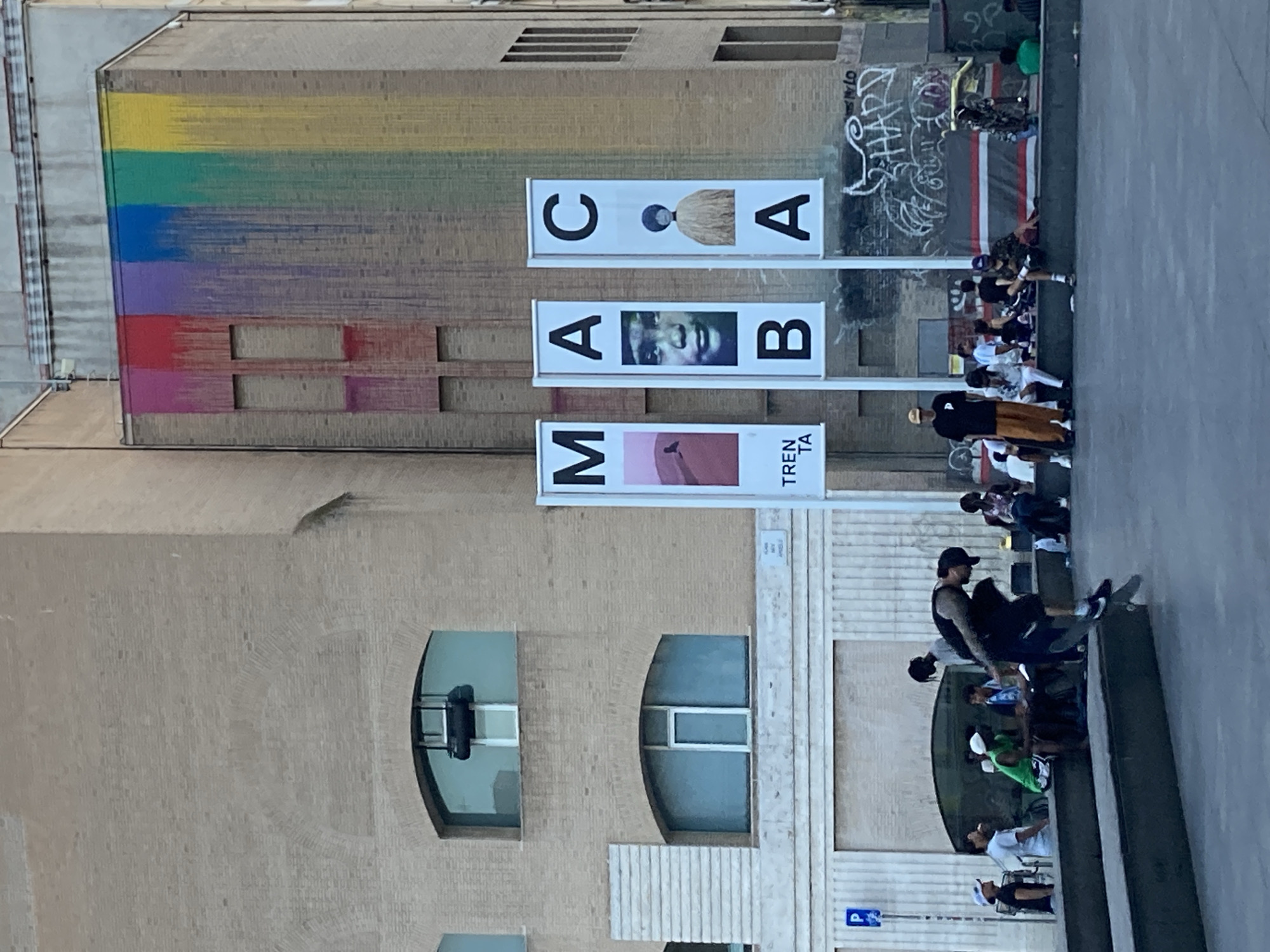 The image depicts an outdoor urban scene with a group of people gathered, some of whom appear to be skateboarding. The setting is a building facade adorned with colorful, striped artwork that resembles a rainbow. Multiple vertical banners with text and images are displayed on the building. One banner shows the letters "C", "A", and "A", with an image of a person in between the first two letters. Another banner displays "A", "A", and "B", with a close-up portrait of a person's face in the middle. A third banner has "M" and "A" on the left, and "TREN TA" on the right, with an image of a person's torso between "A" and "TREN".
Several people are visible in the foreground and middle ground. Some are sitting, while others are standing or engaged in activities. One person in the foreground is on a skateboard, wearing a black tank top and a hat. Others are dressed casually. The building itself is constructed of light-colored bricks and features rounded windows. Graffiti is visible on a section of the wall behind the banners.
The scene appears to be outdoors during daylight, as indicated by the lighting. The atmosphere seems casual and relaxed, with a focus on the people and the art. While specific time of day and weather are not explicitly stated, the bright lighting suggests a sunny day. The presence of graffiti and street art, along with the casual gathering of people, hints at a vibrant urban environment. The text on the banners suggests that the location might be related to an exhibition or event.